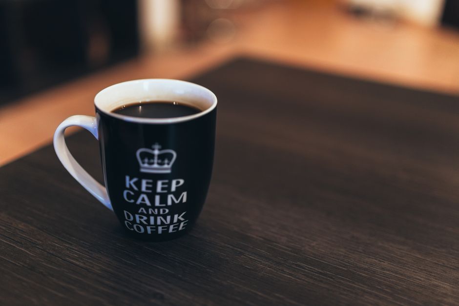 Black mug with 'Keep Calm and Drink Coffee' text, sitting on a dark wooden table.