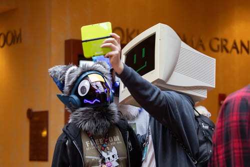 A person with a computer outfited as a mask taking a selfie with a fursuiter using a DS.