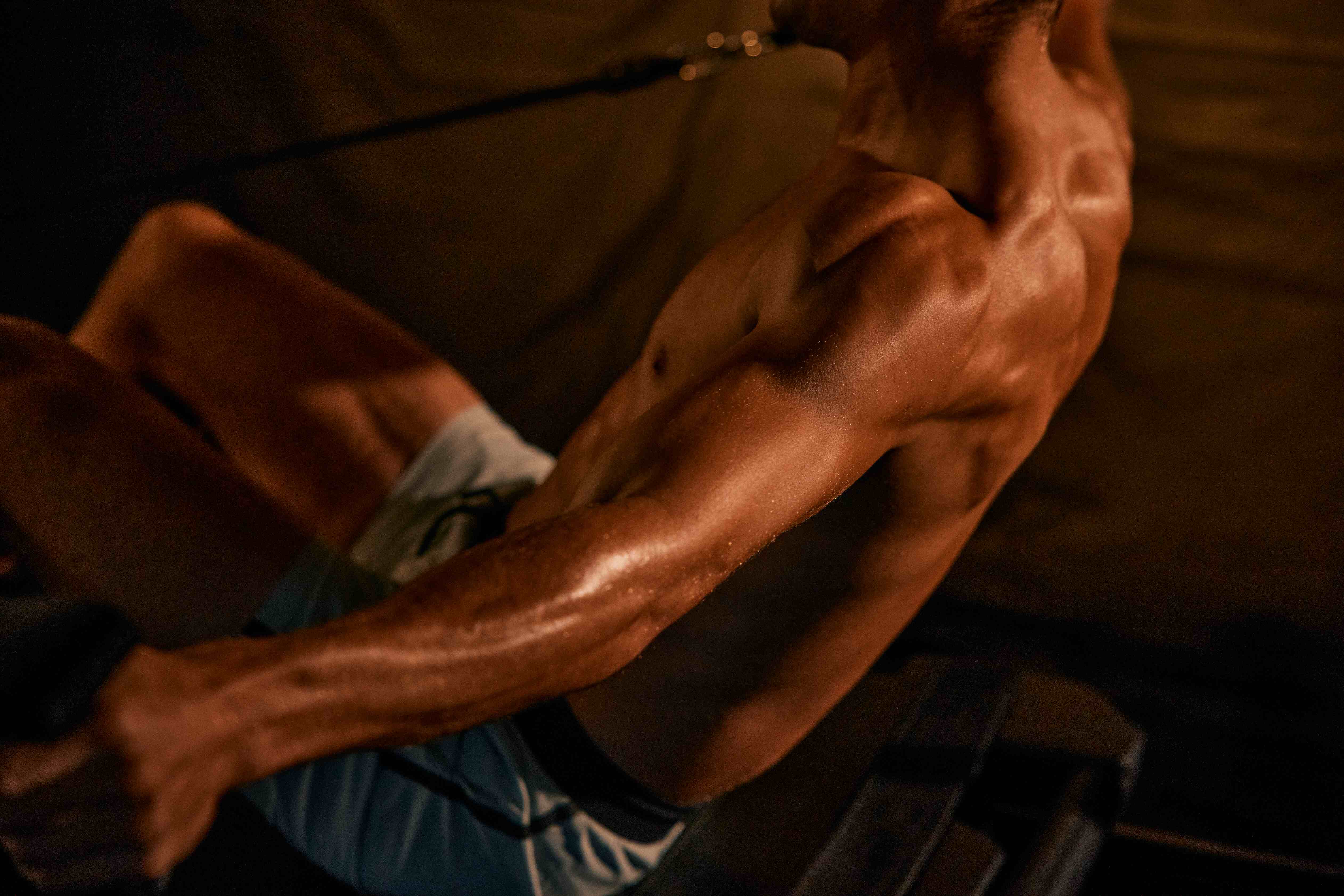 Muscular shirtless man with defined arm and shoulder muscles working out on exercise equipment in dim lighting.
