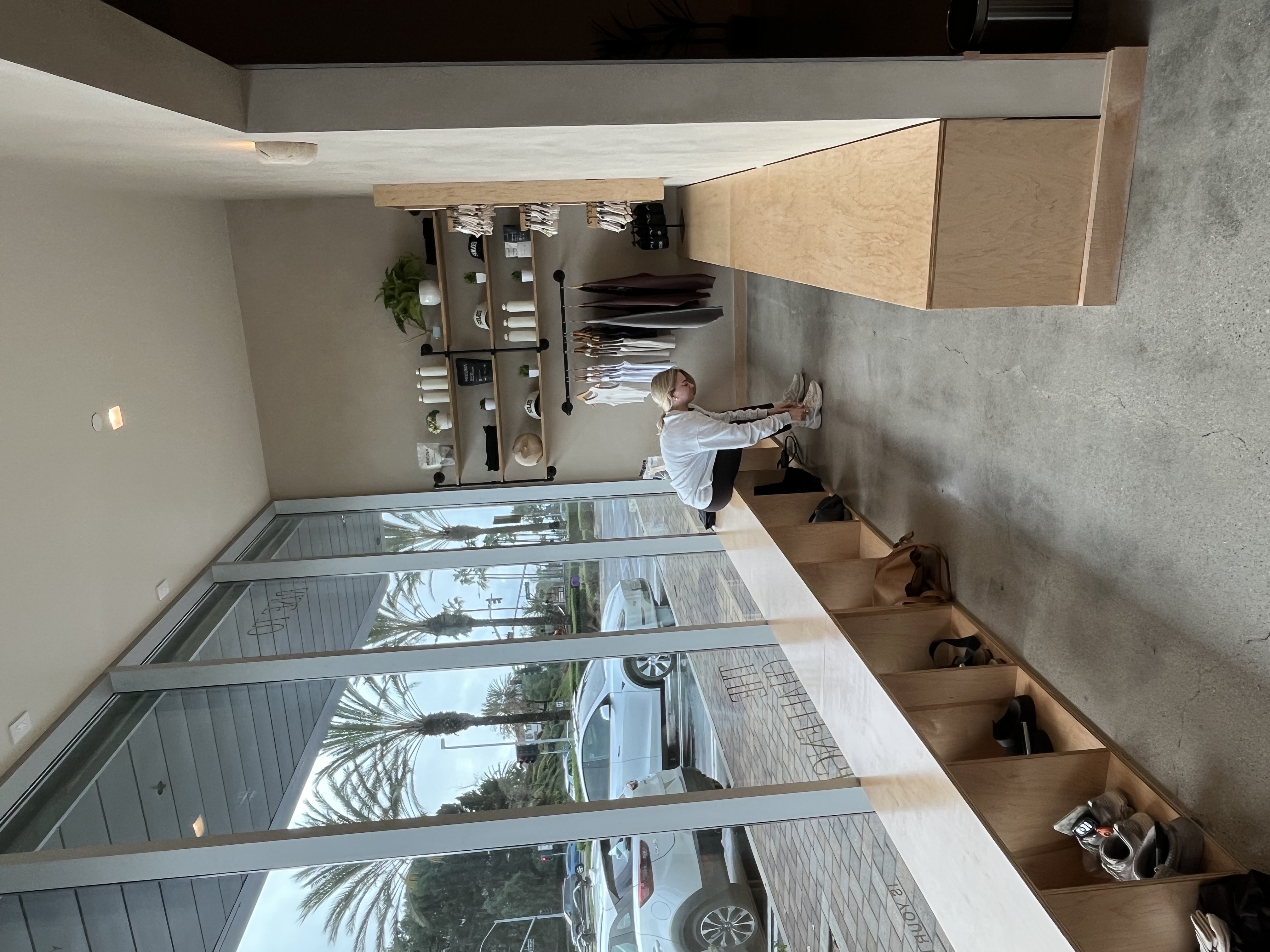 Woman sitting on wooden bench in a modern store, tying her shoes, with shelves and clothing displayed on the wall beside her.