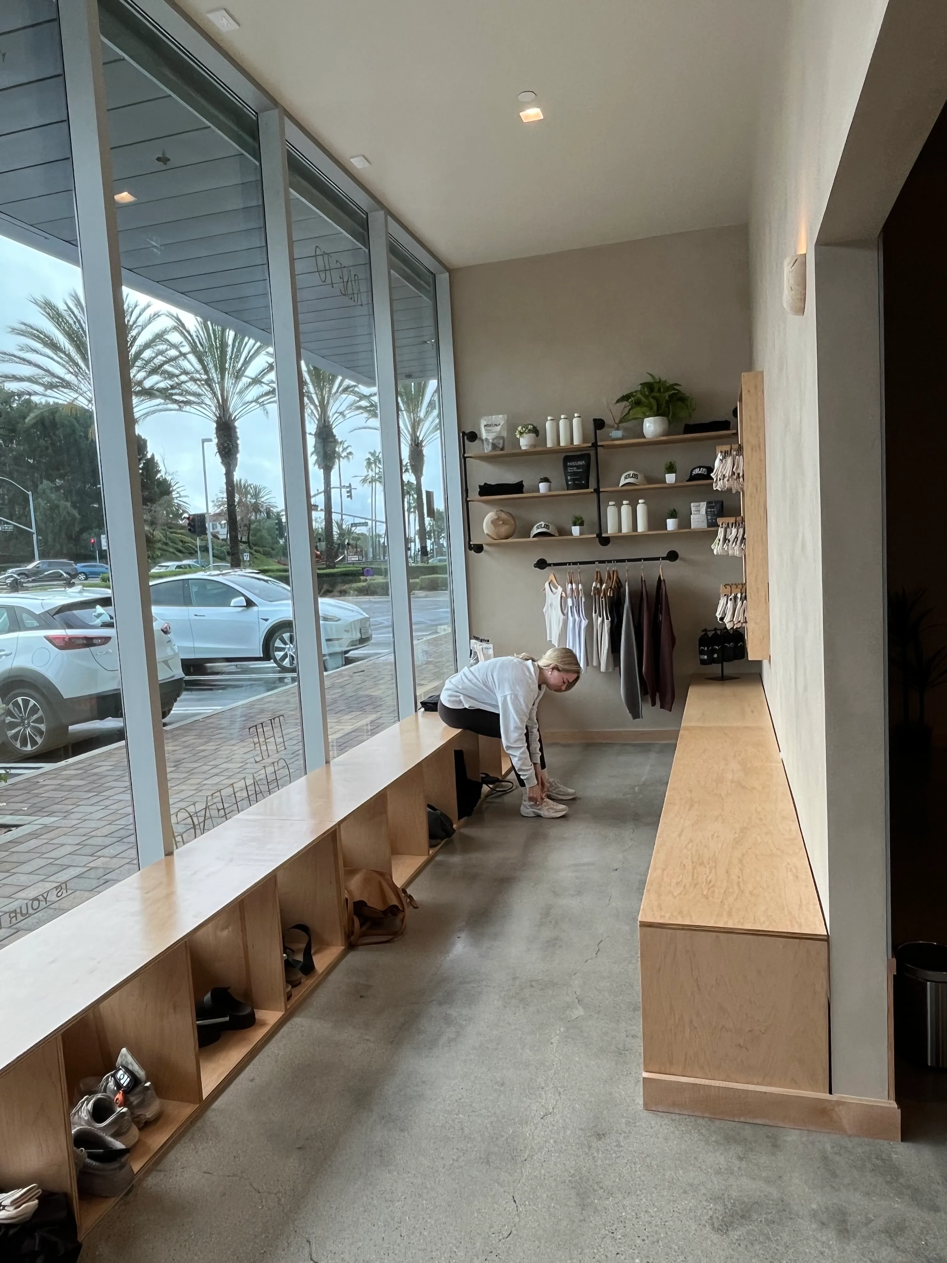 Woman sitting on wooden bench in a modern store, tying her shoes, with shelves and clothing displayed on the wall beside her.