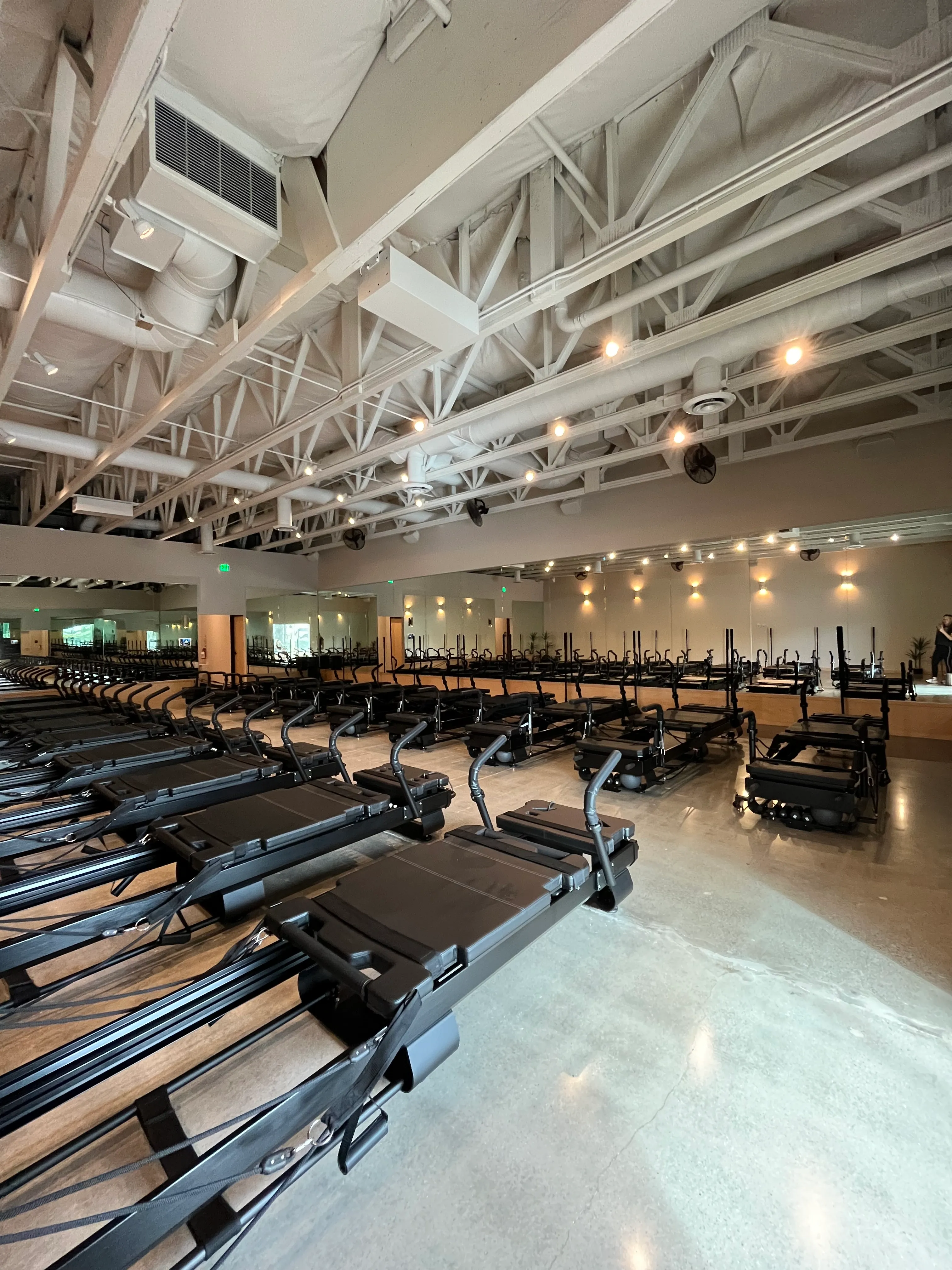 Spacious Pilates studio with rows of black reformer machines on polished concrete floor under exposed ceiling with lighting.