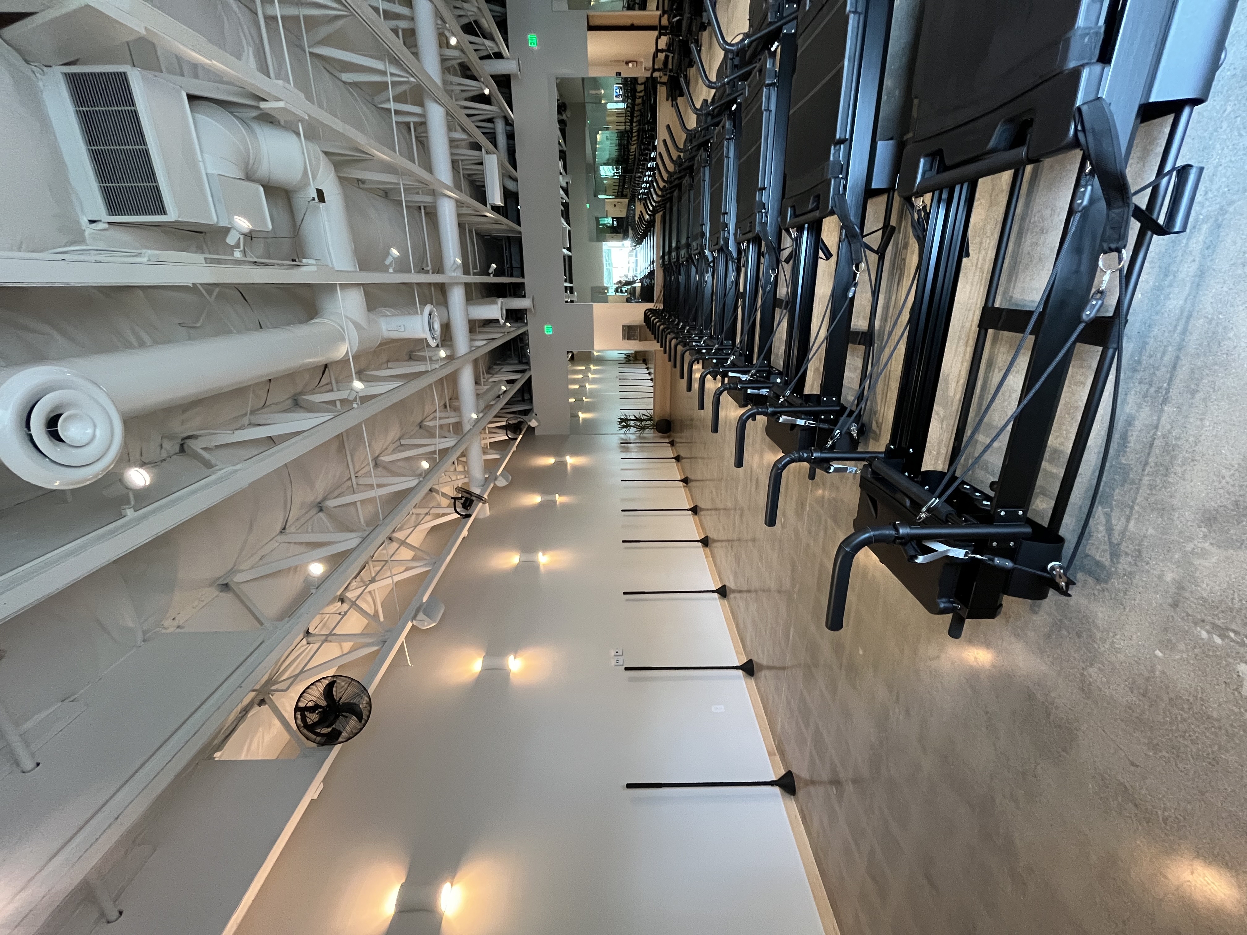 Interior view of a modern Pilates studio with a row of black reformer machines on the right and wall bars on the left.