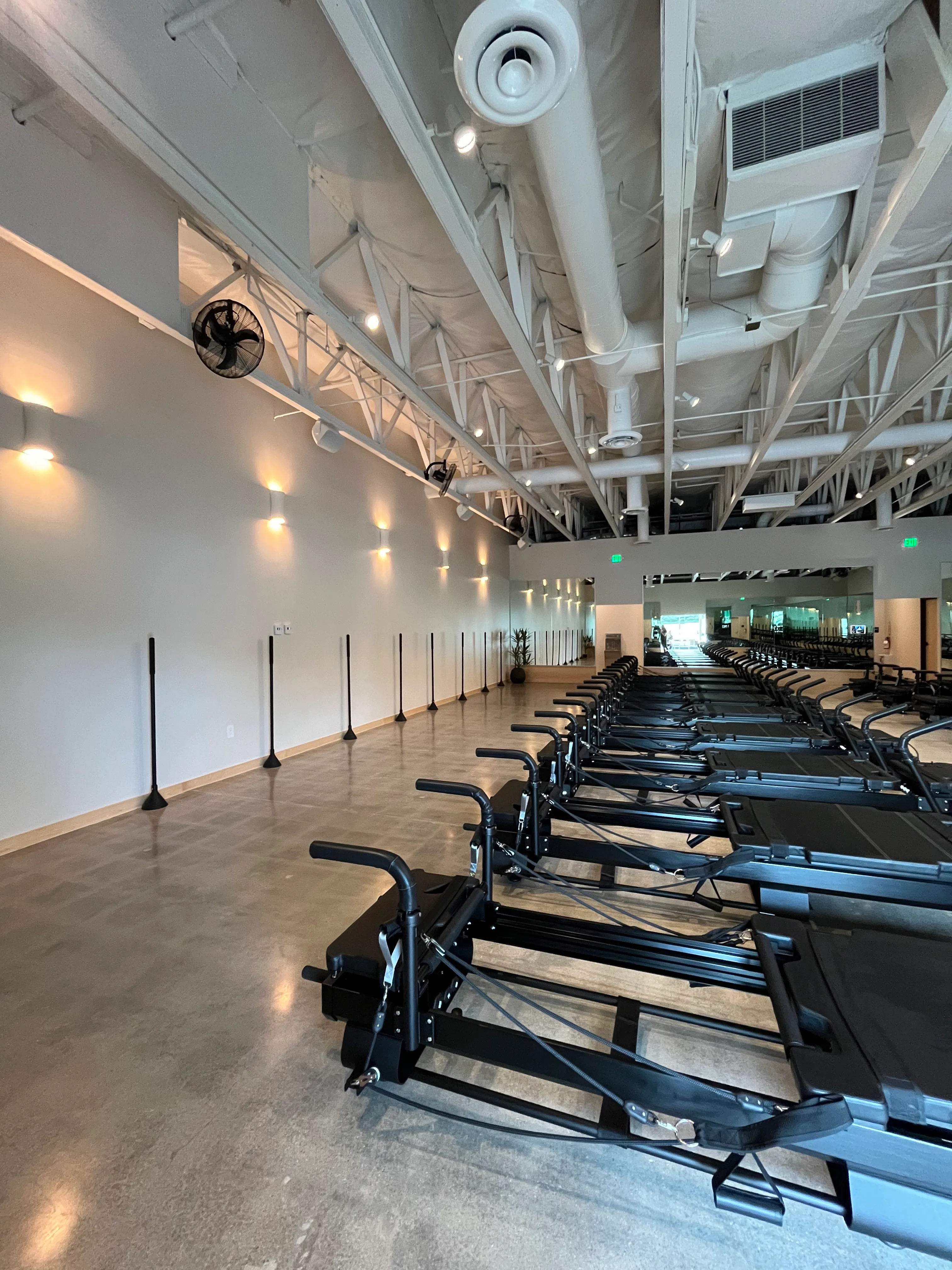 Interior view of a modern Pilates studio with a row of black reformer machines on the right and wall bars on the left.