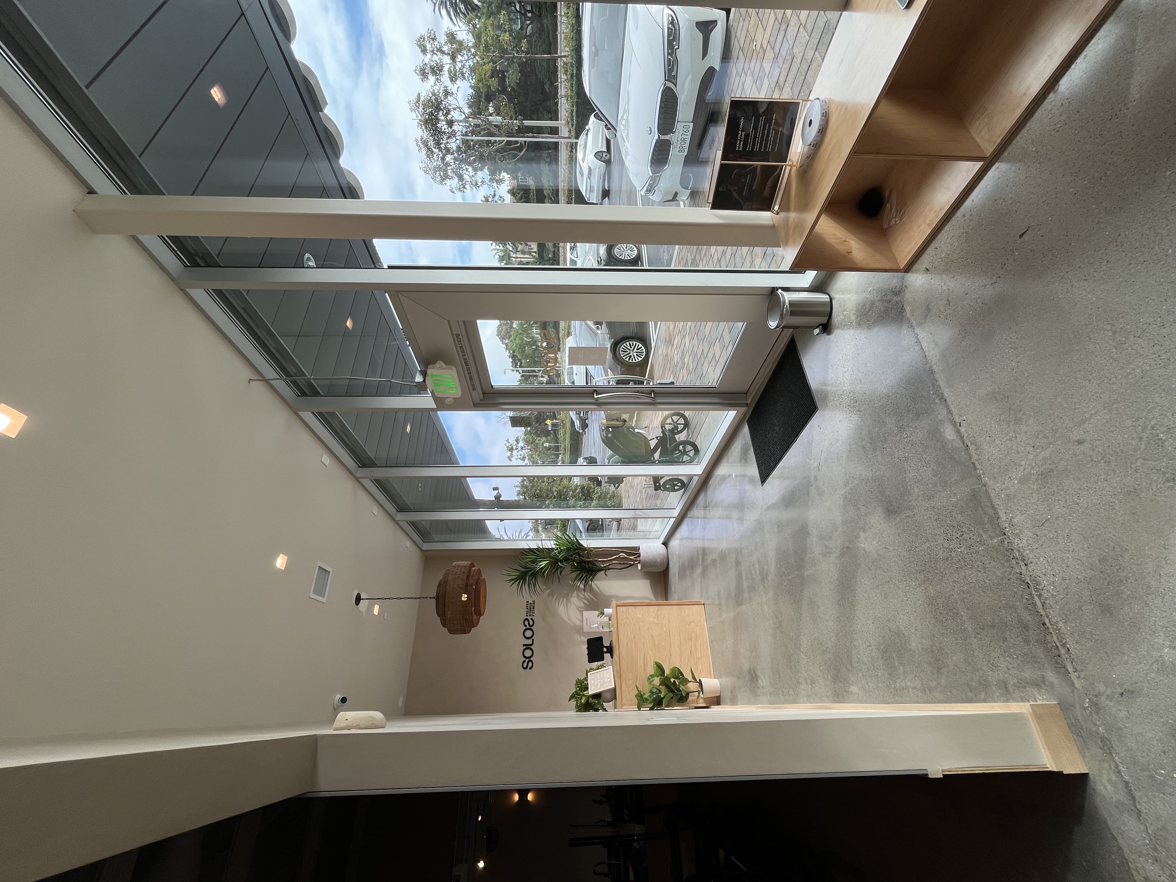 Interior view of a modern studio entrance with glass walls, wooden reception desk, plants, and parked cars visible outside.