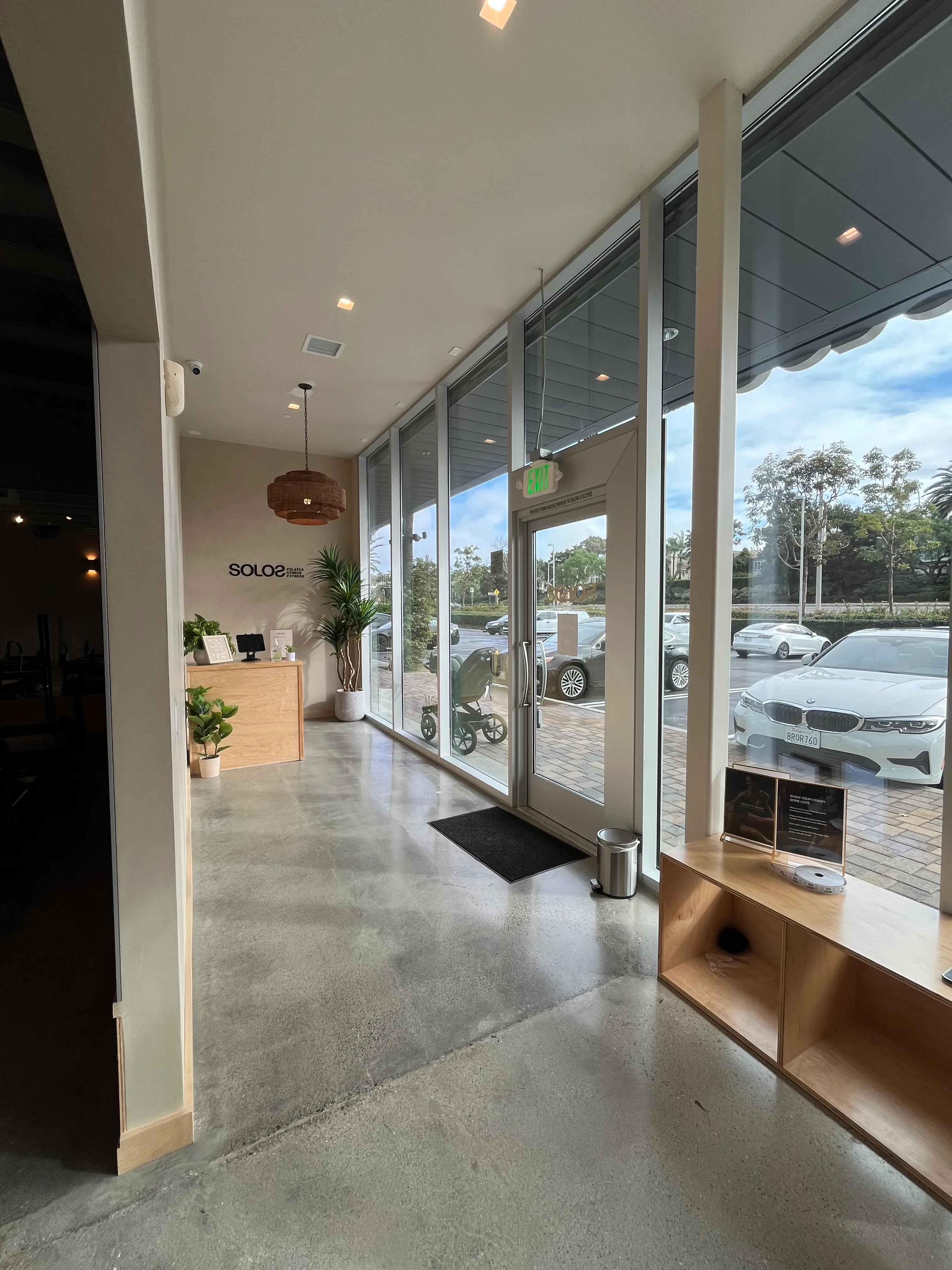 Interior view of a modern studio entrance with glass walls, wooden reception desk, plants, and parked cars visible outside.