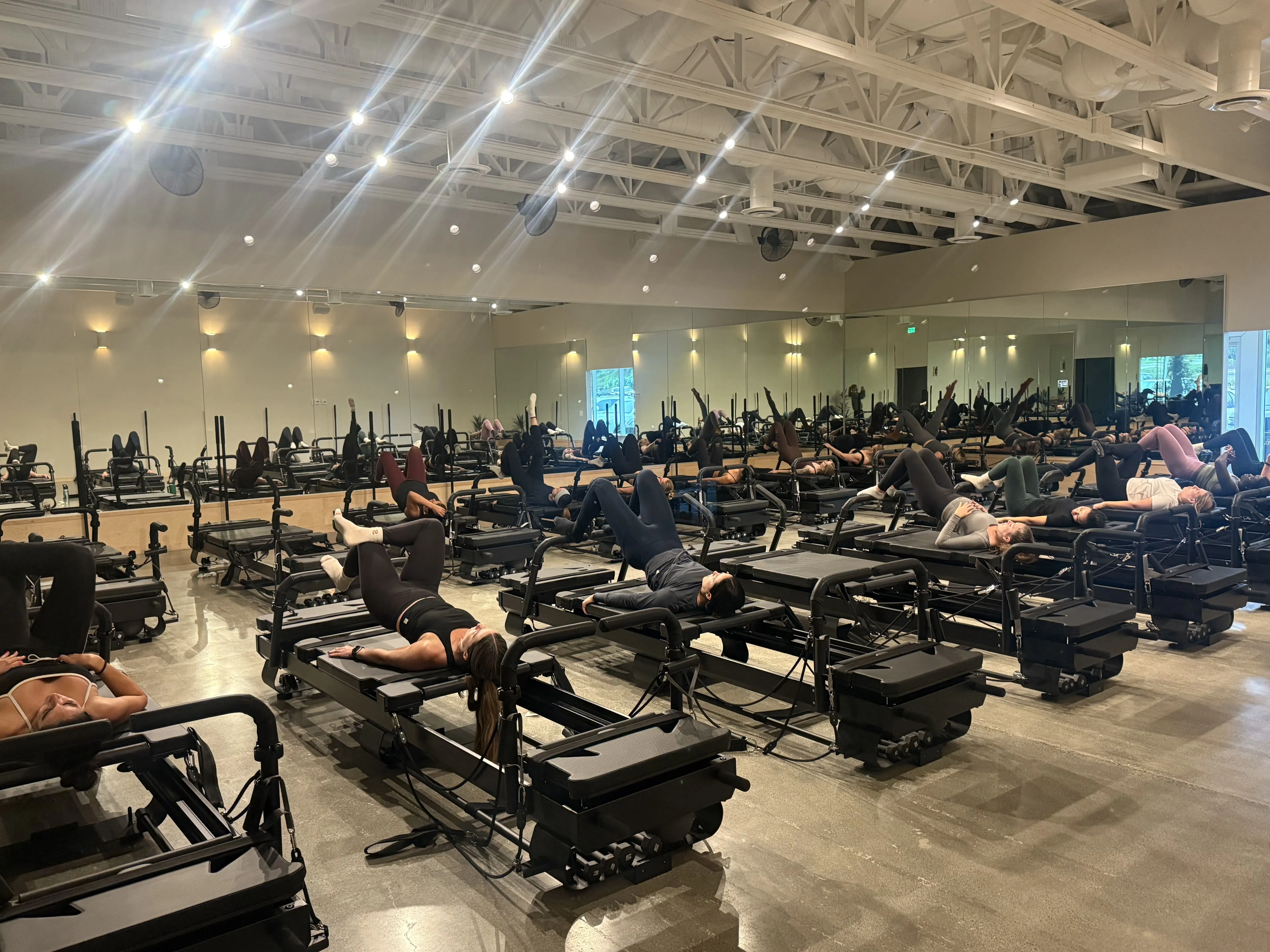 People exercising on Pilates reformer machines in a large studio with mirrored walls and bright ceiling lights.