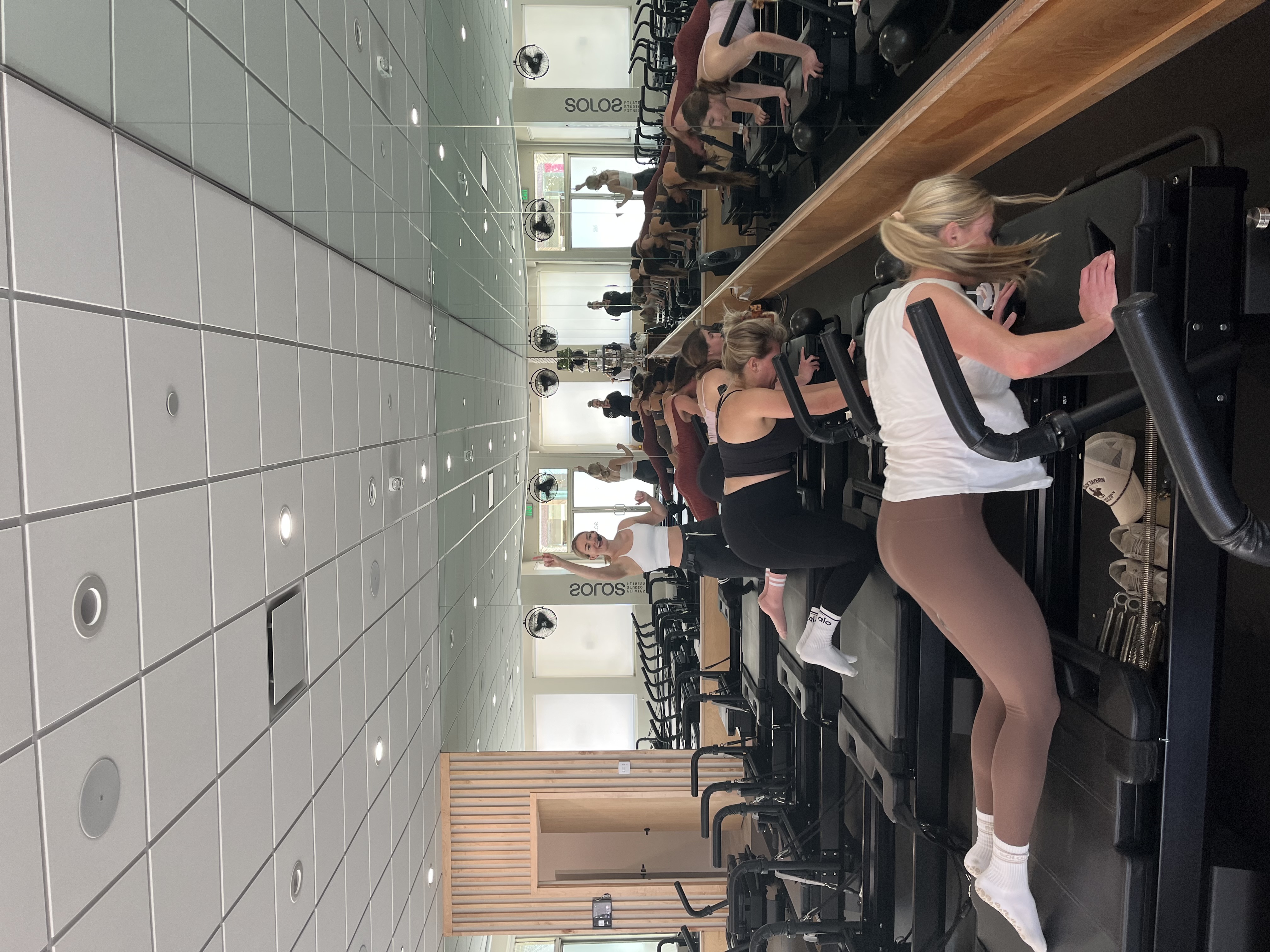 Group fitness class with women performing exercises on Pilates reformer machines in front of a large mirror.