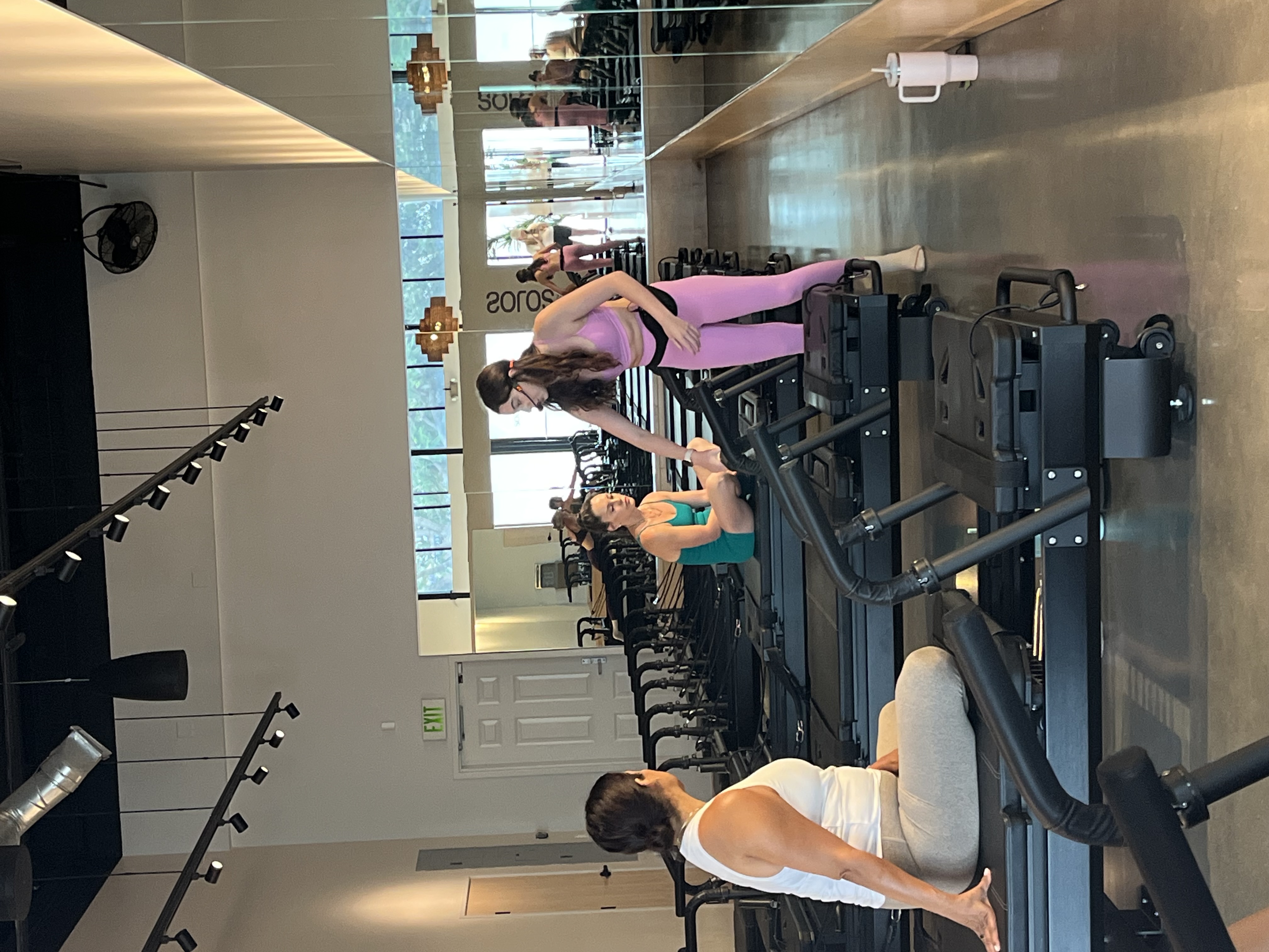 Fitness instructor in pink workout clothes assisting a woman in teal seated on a Pilates reformer machine inside a gym studio.