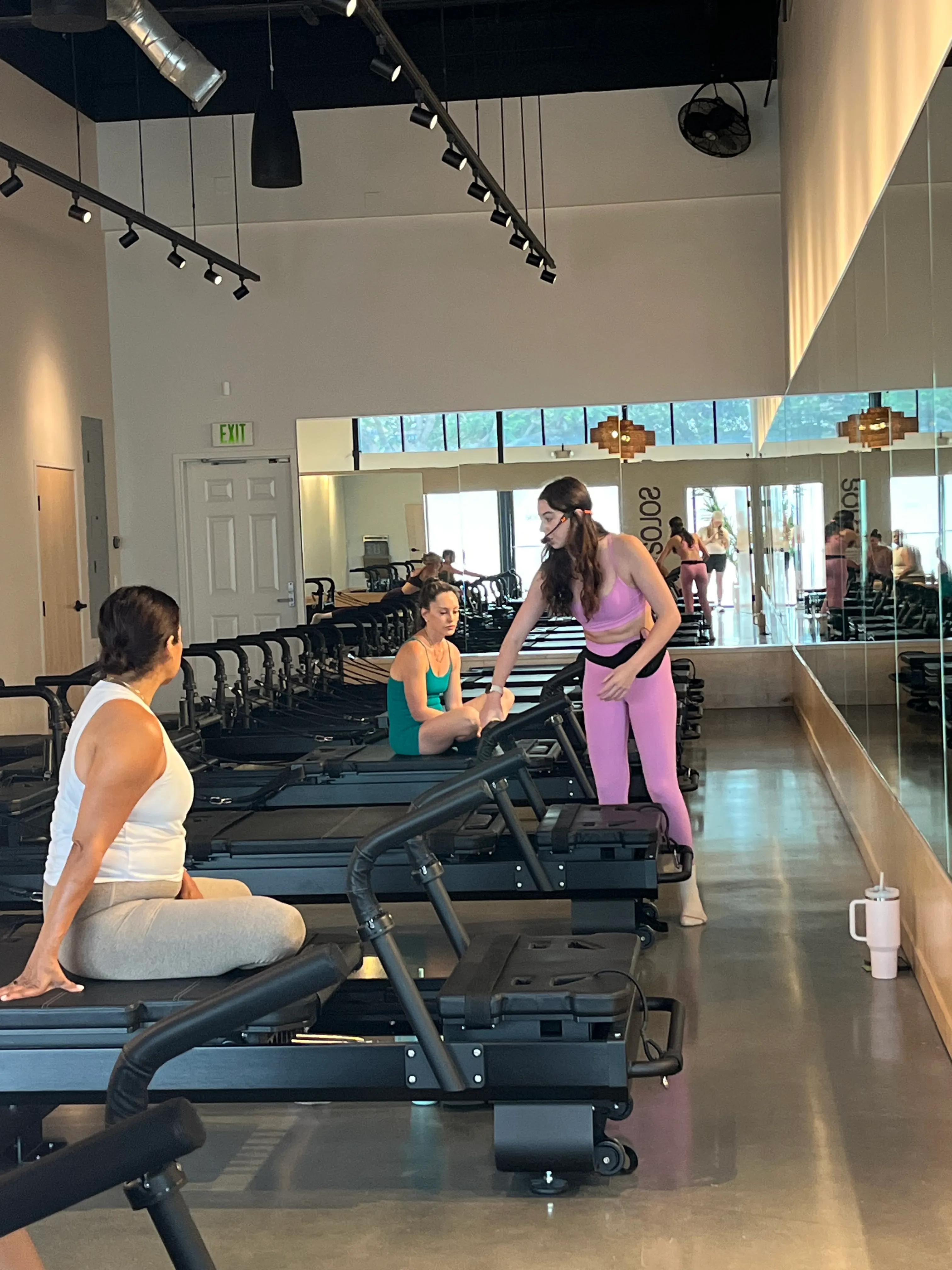 Fitness instructor in pink workout clothes assisting a woman in teal seated on a Pilates reformer machine inside a gym studio.