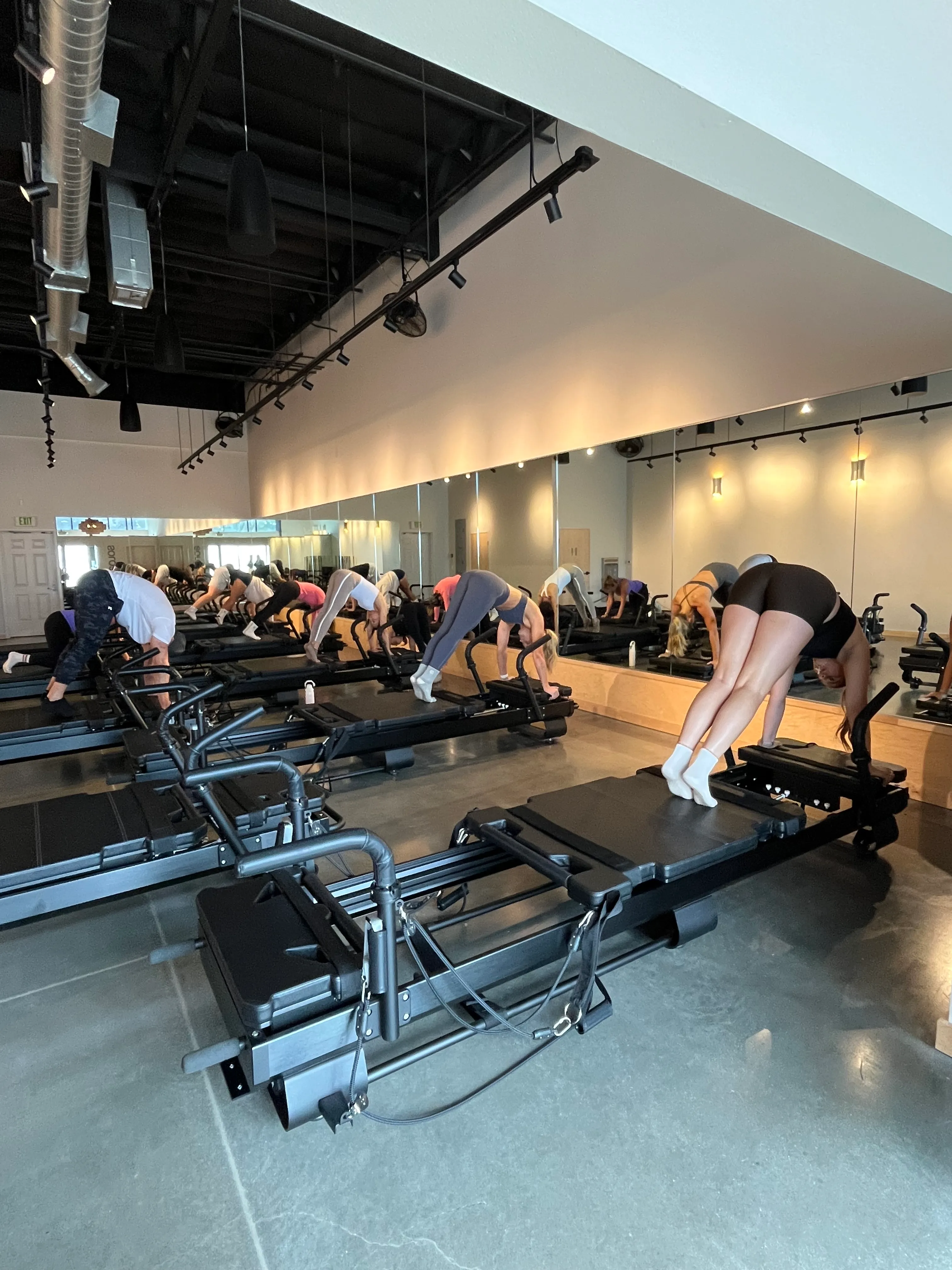 A group doing Pilates on reformer machines in a mirrored studio with polished concrete floor and exposed ceiling.