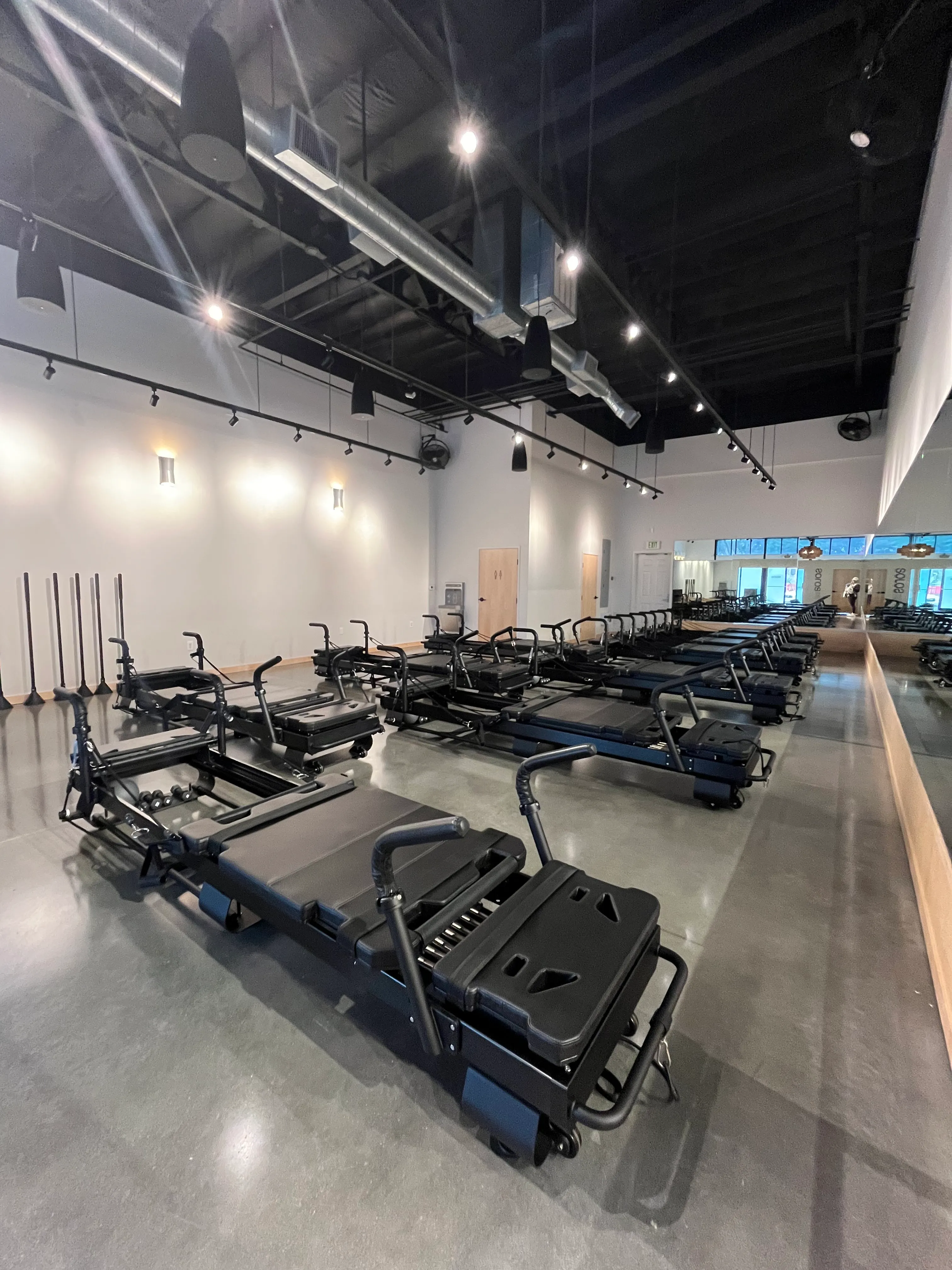 Interior of a modern Pilates studio with multiple reformer machines arranged in rows on a polished concrete floor, large mirror on one wall, and industrial ceiling with lights and ducts.