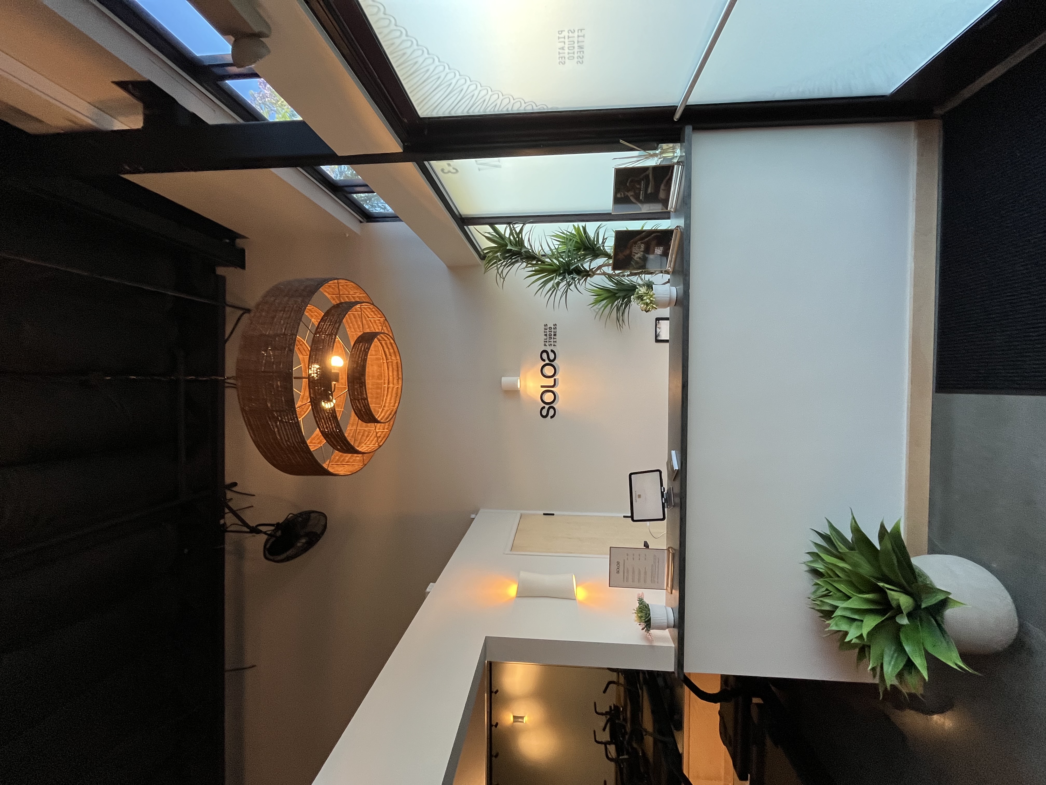 Reception area of a Pilates studio with a white counter, green plants, a circular hanging light, and 'SOLOS Pilates Studio Fitness' sign on the wall.