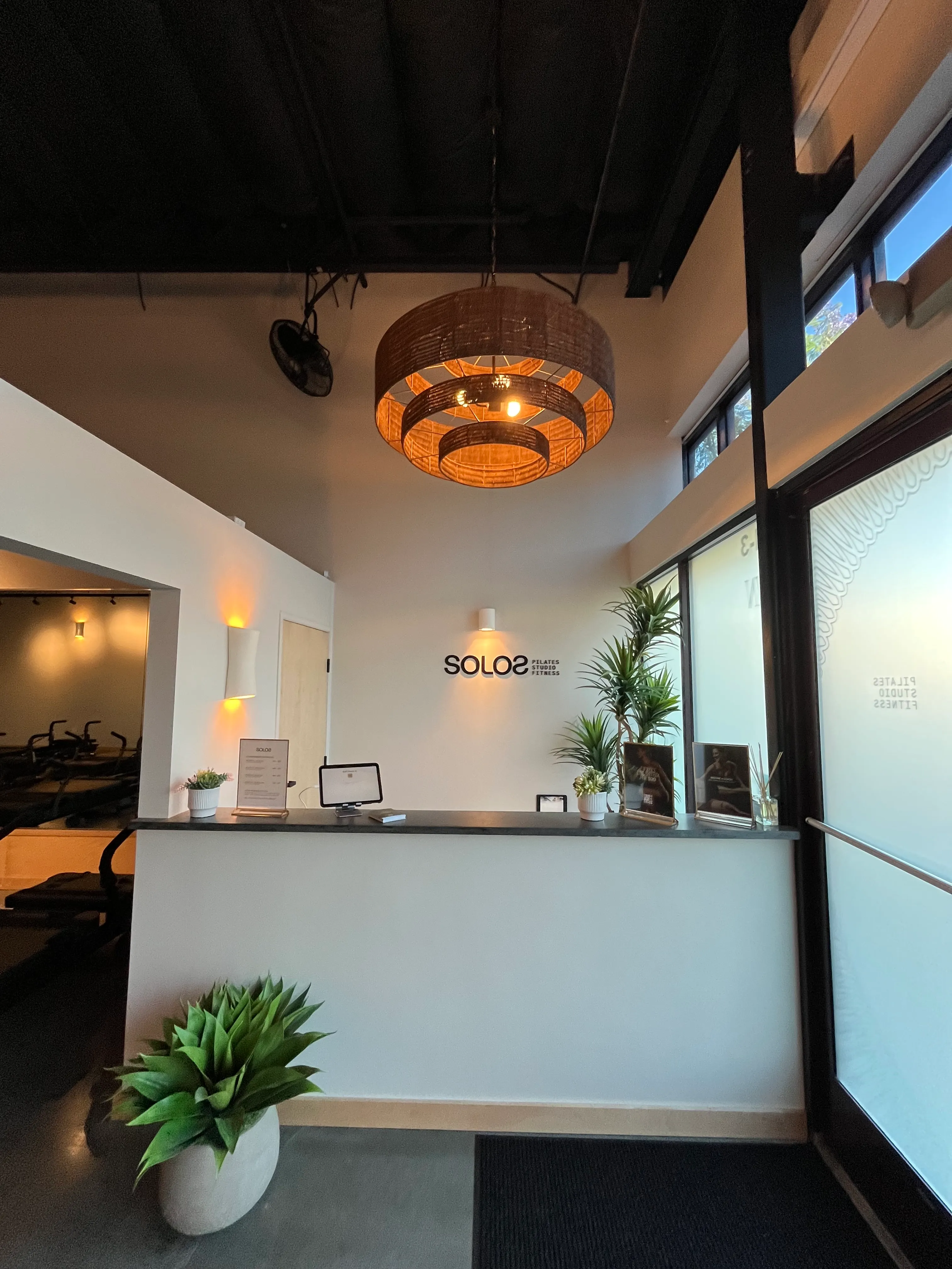 Reception area of a Pilates studio with a white counter, green plants, a circular hanging light, and 'SOLOS Pilates Studio Fitness' sign on the wall.