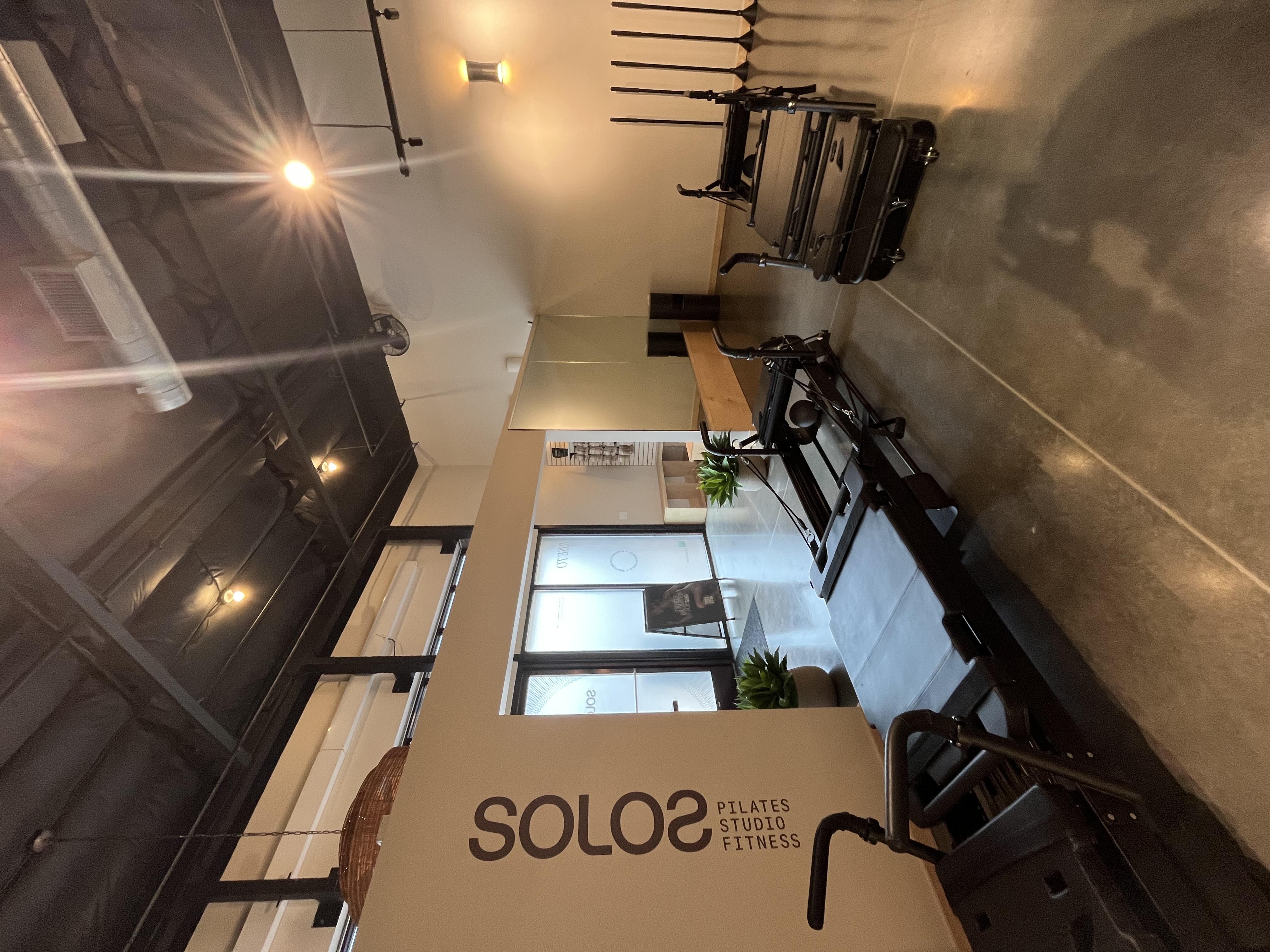 Indoor Pilates studio with reformer machines, polished concrete floors, and light beige walls with the sign 'SOLOS Pilates Studio Fitness'.