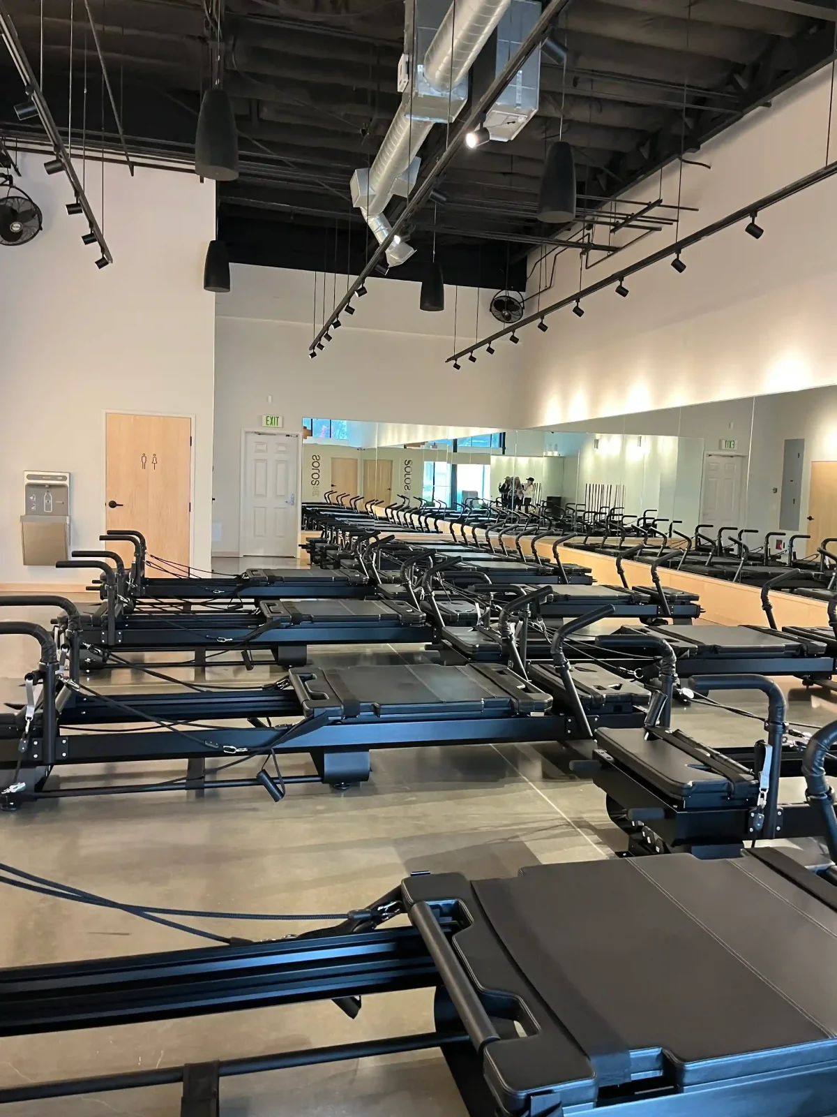 Indoor Pilates studio with multiple black reformer machines arranged in rows on a polished concrete floor and mirrors on one wall.