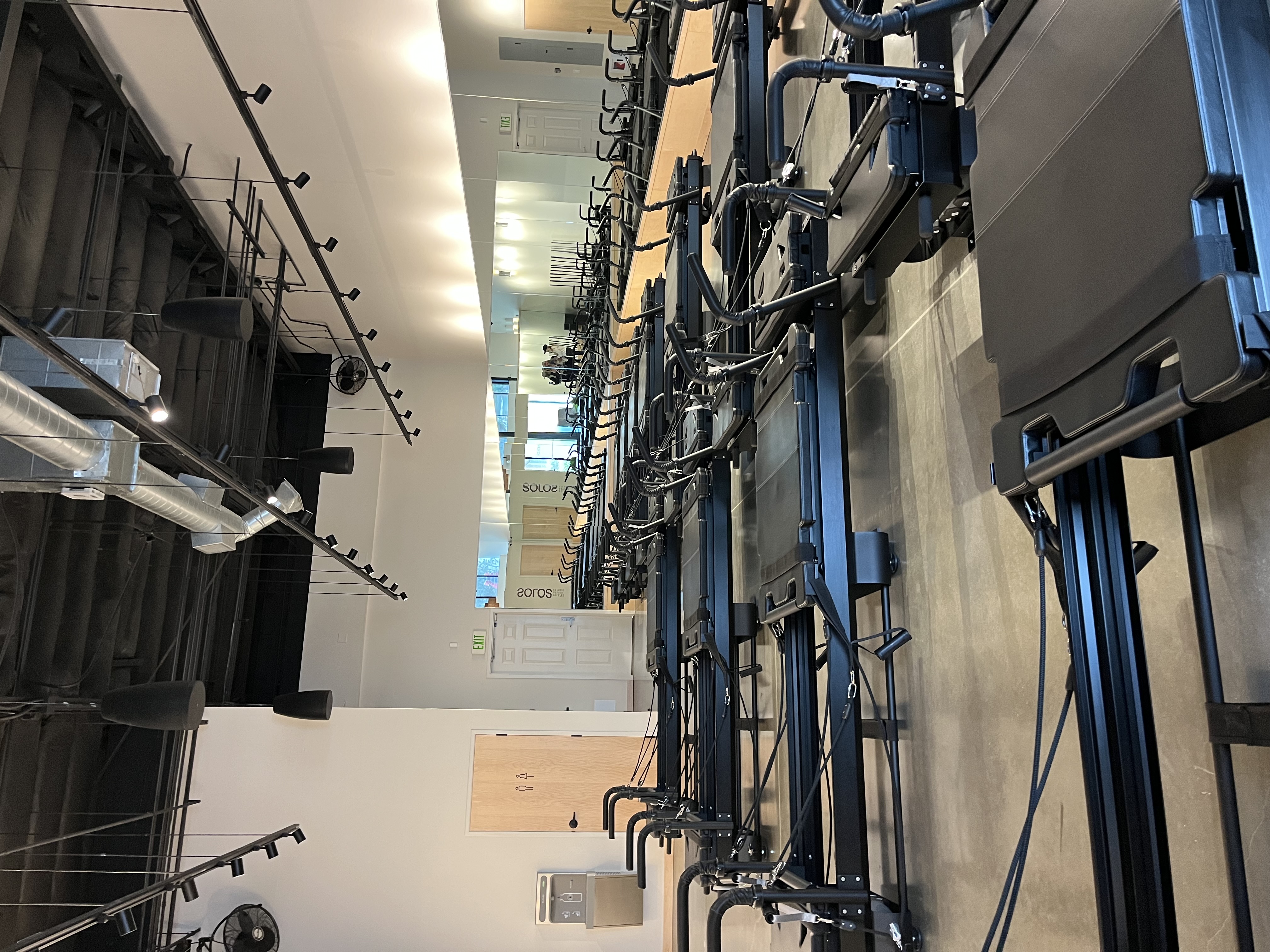 Indoor Pilates studio with multiple black reformer machines arranged in rows on a polished concrete floor and mirrors on one wall.