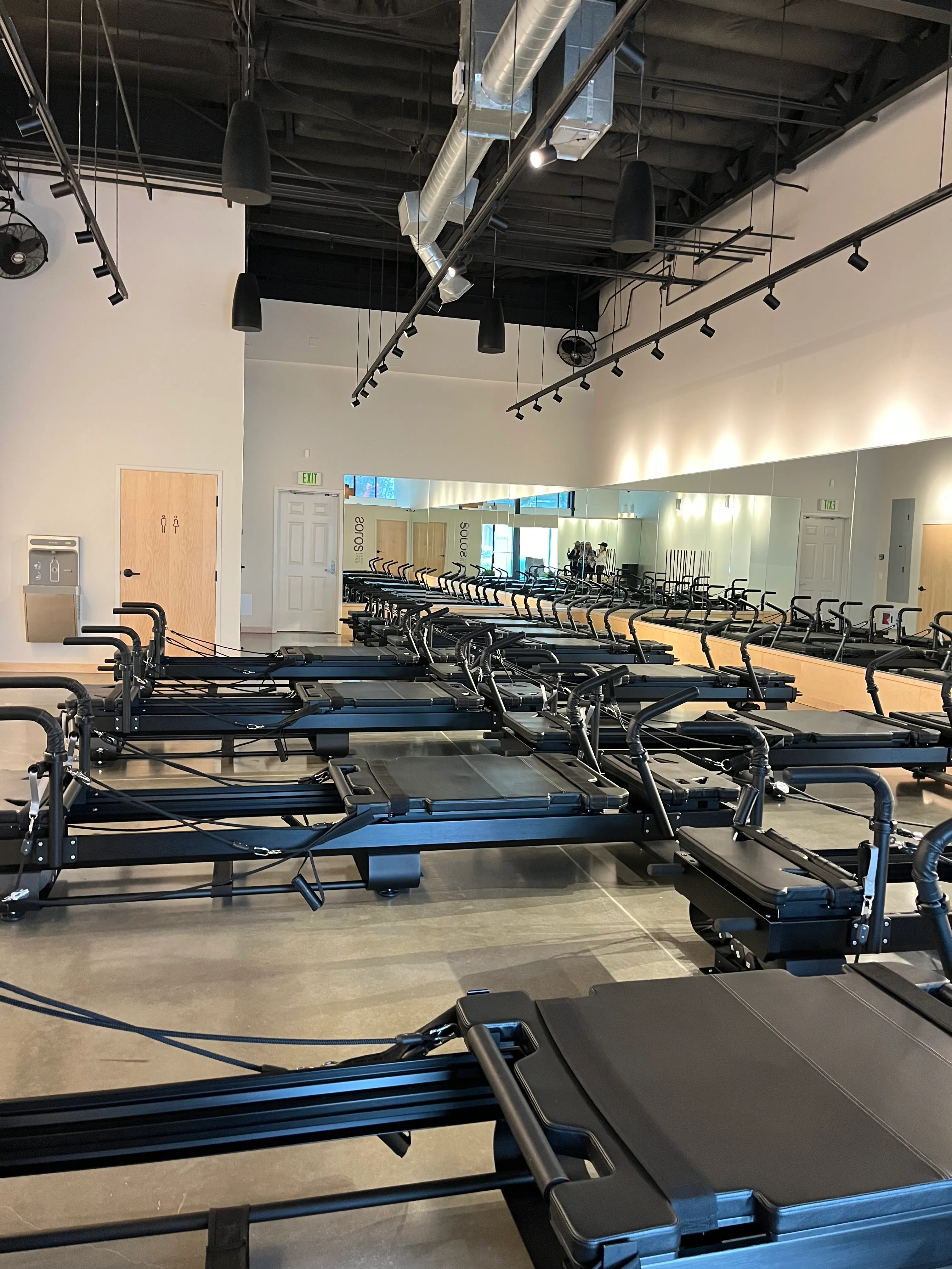 Indoor Pilates studio with multiple black reformer machines arranged in rows on a polished concrete floor and mirrors on one wall.