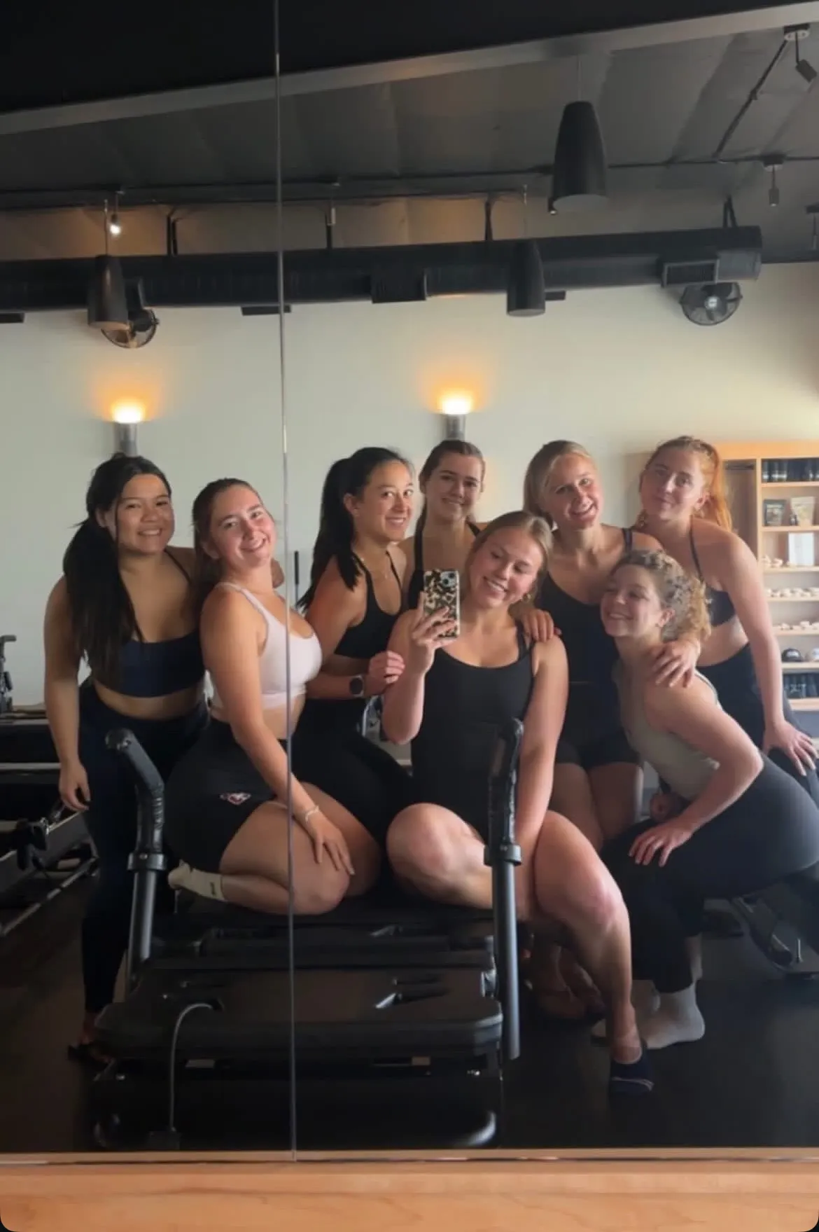 Group of 12 women posing in a fitness studio with pilates reformer machines and mirrors in the background.