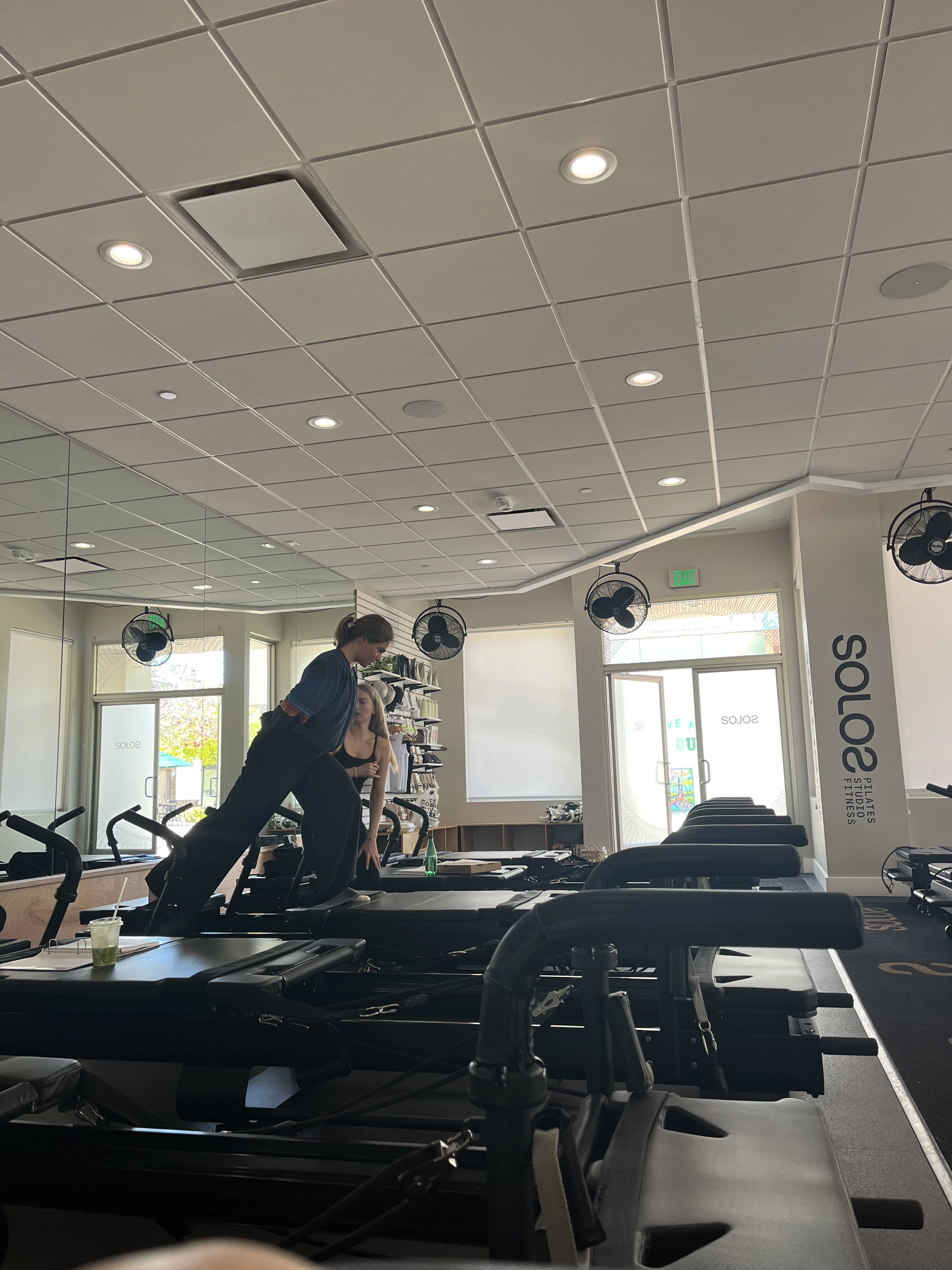 Two women in a Pilates studio, one guiding the other on a reformer machine amid workout equipment and large windows.