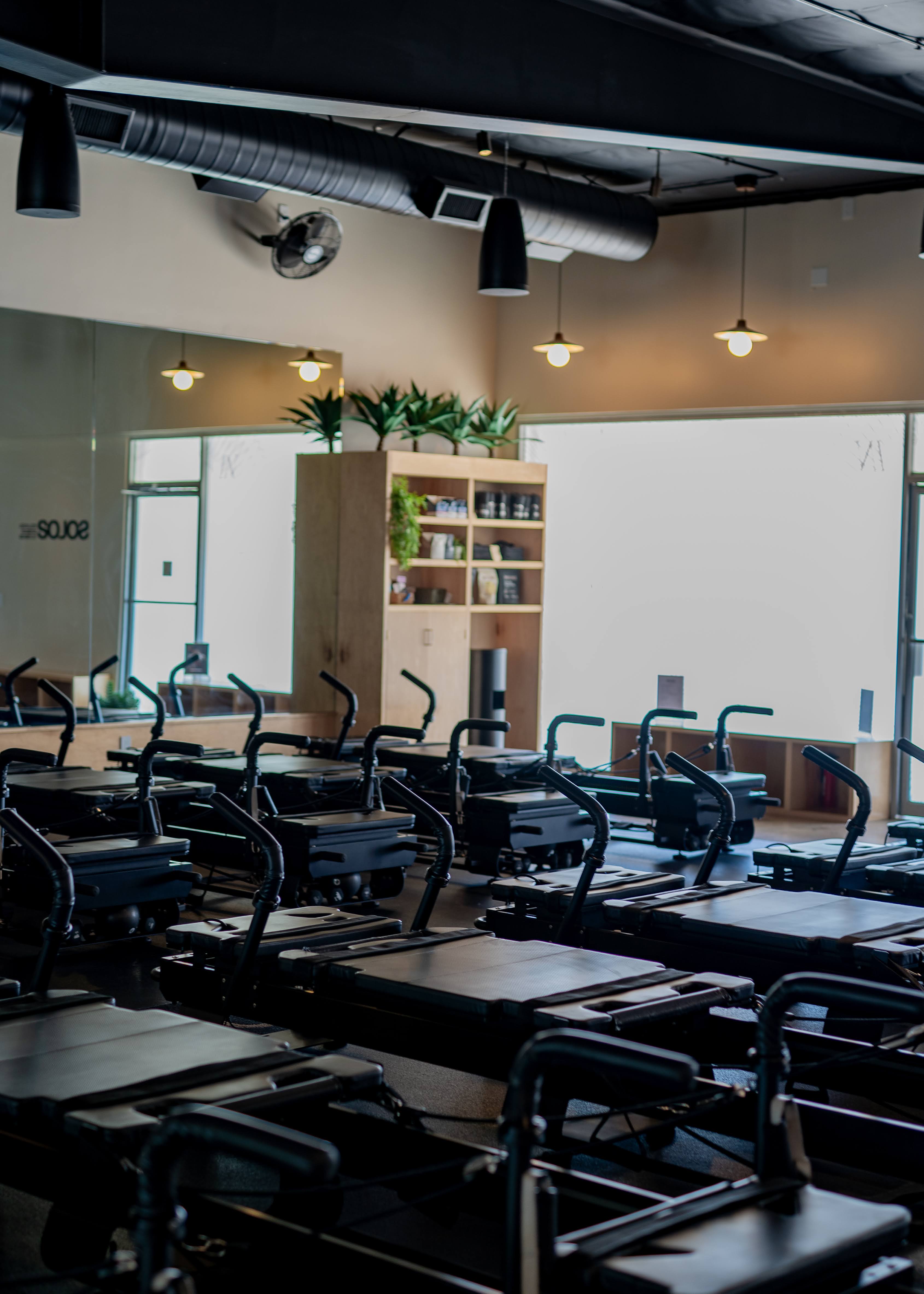 Empty Pilates reformer machines neatly arranged in a bright, modern studio with wooden shelves and plants in the background.