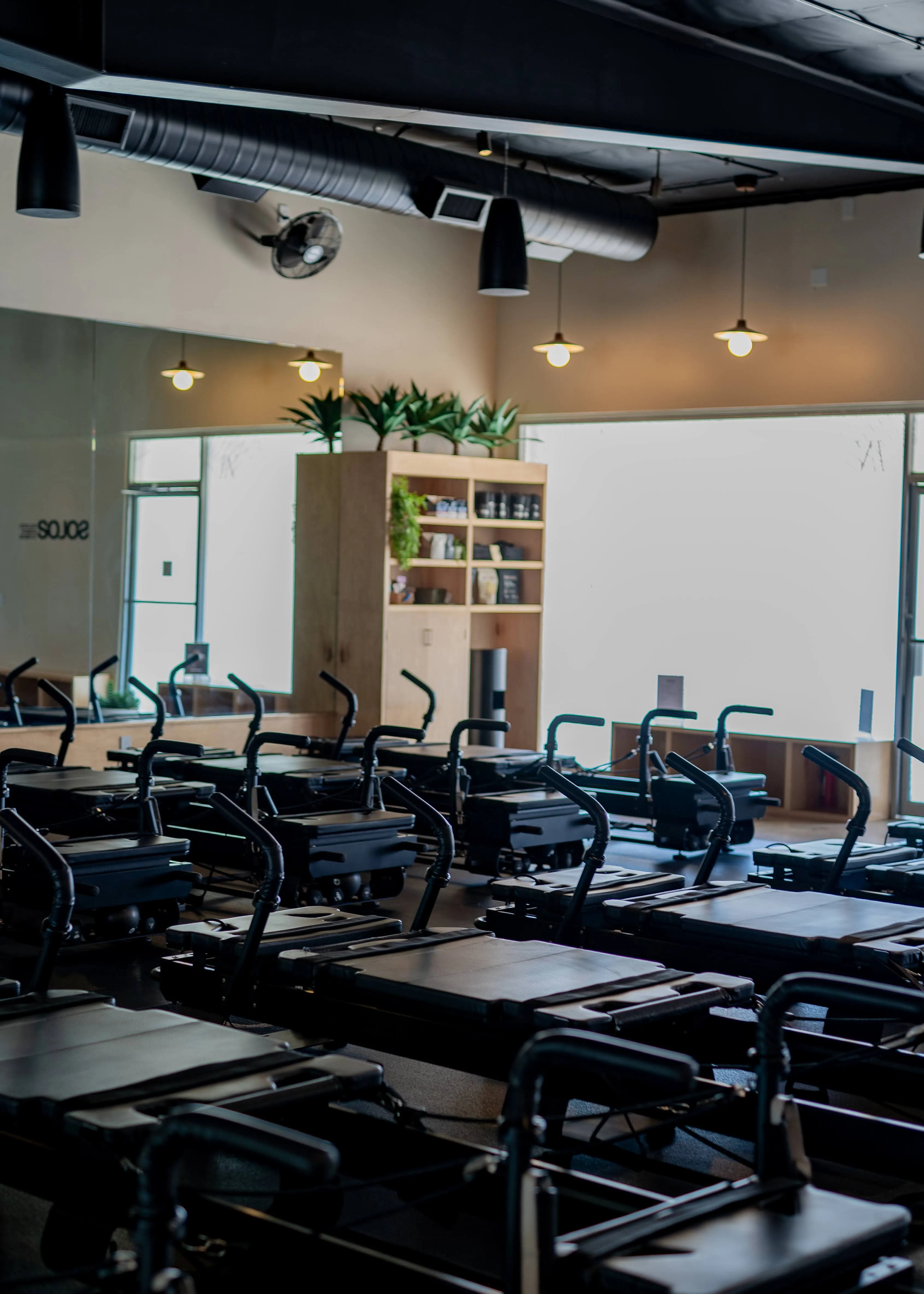 Empty Pilates reformer machines neatly arranged in a bright, modern studio with wooden shelves and plants in the background.