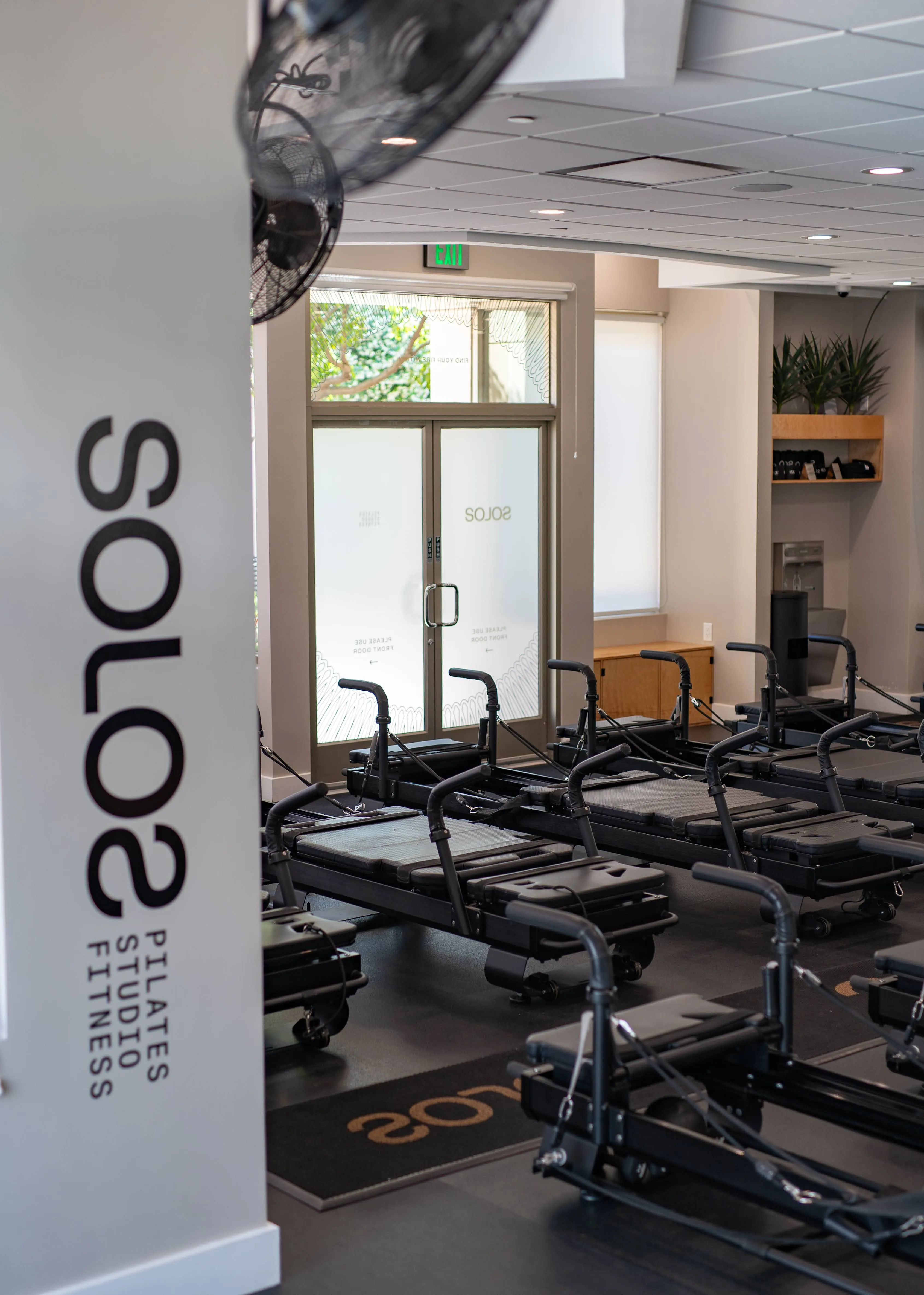 Interior of a Pilates studio featuring multiple black reformer machines arranged in rows and a glass door with the word 'SOLOS'.
