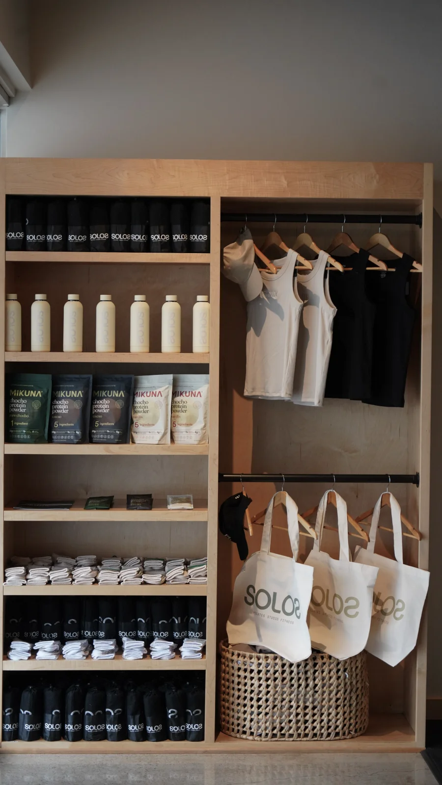 Wooden shelving unit with black SOLO2 towels, white Solo2 bottles, Mikuna protein powder packets, folded towels, white and black tank tops, beige caps, and white SOLO2 tote bags.