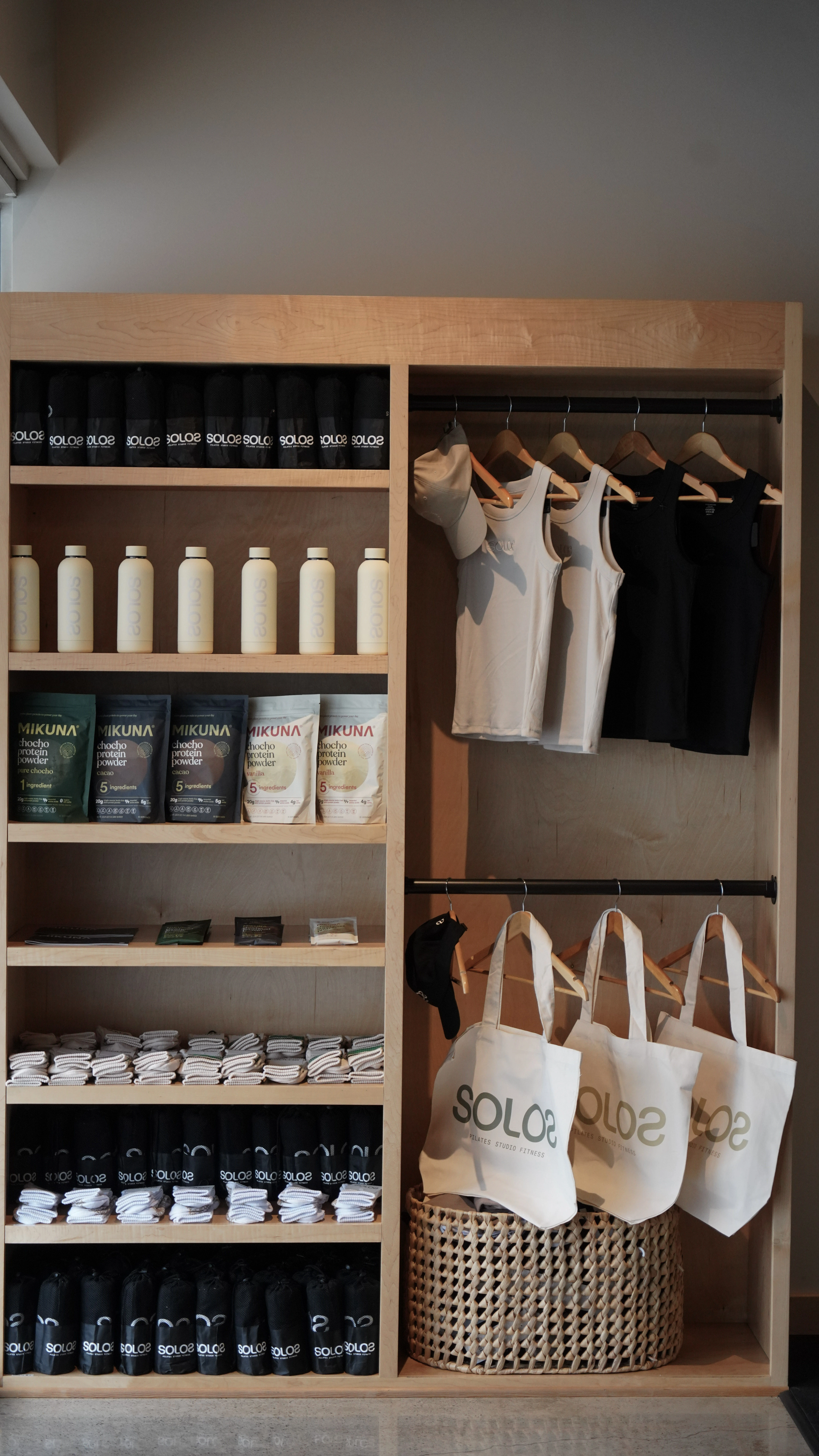 Wooden shelving unit with black SOLO2 towels, white Solo2 bottles, Mikuna protein powder packets, folded towels, white and black tank tops, beige caps, and white SOLO2 tote bags.