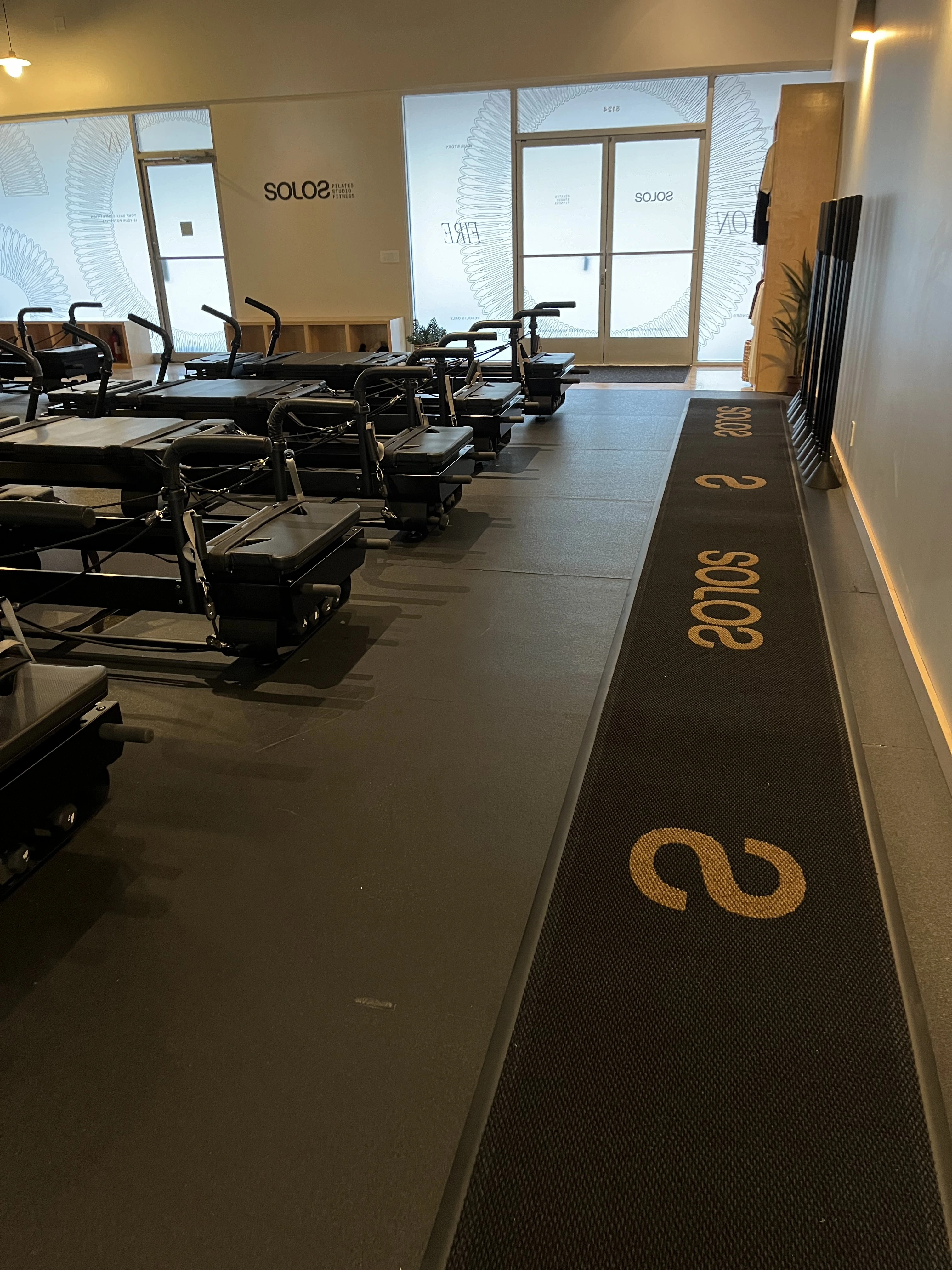 Interior of a Pilates studio with reformer machines and a black mat with gold 'SOLOS' text.