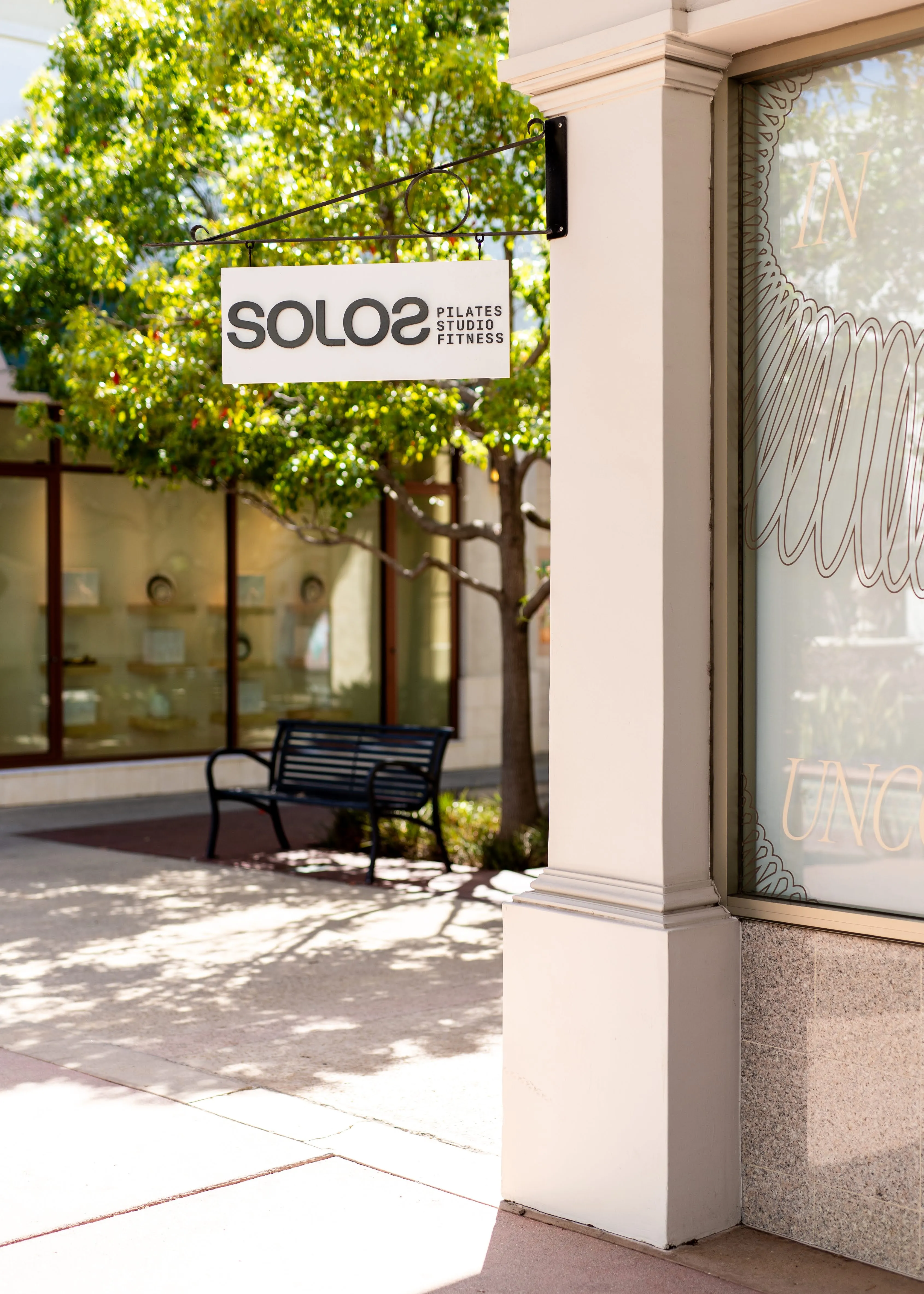 Outdoor sign for Solos Pilates Studio Fitness hangs near a bench and tree by a building.