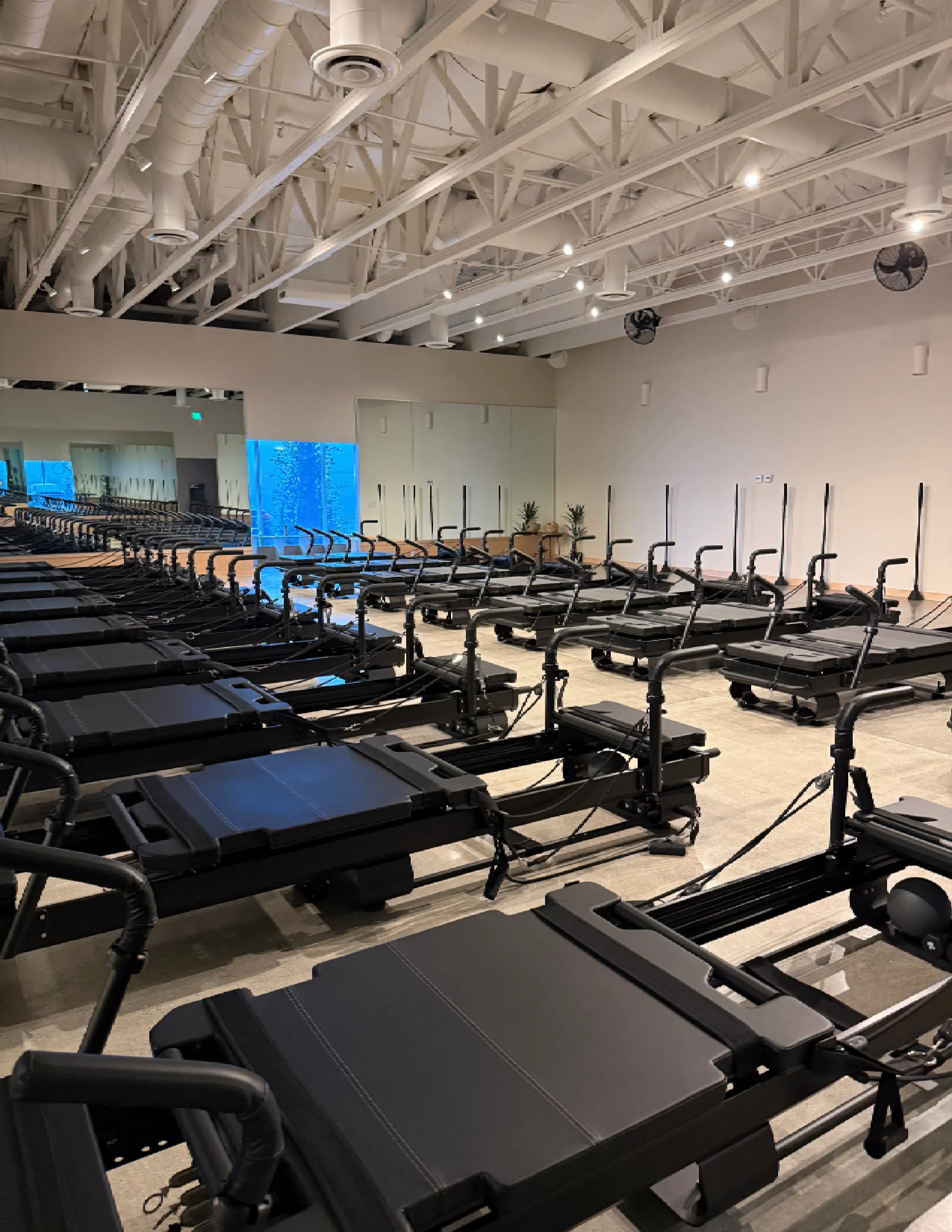 Row of black Pilates reformer machines in a spacious, well-lit fitness studio with high ceiling and mirrors.