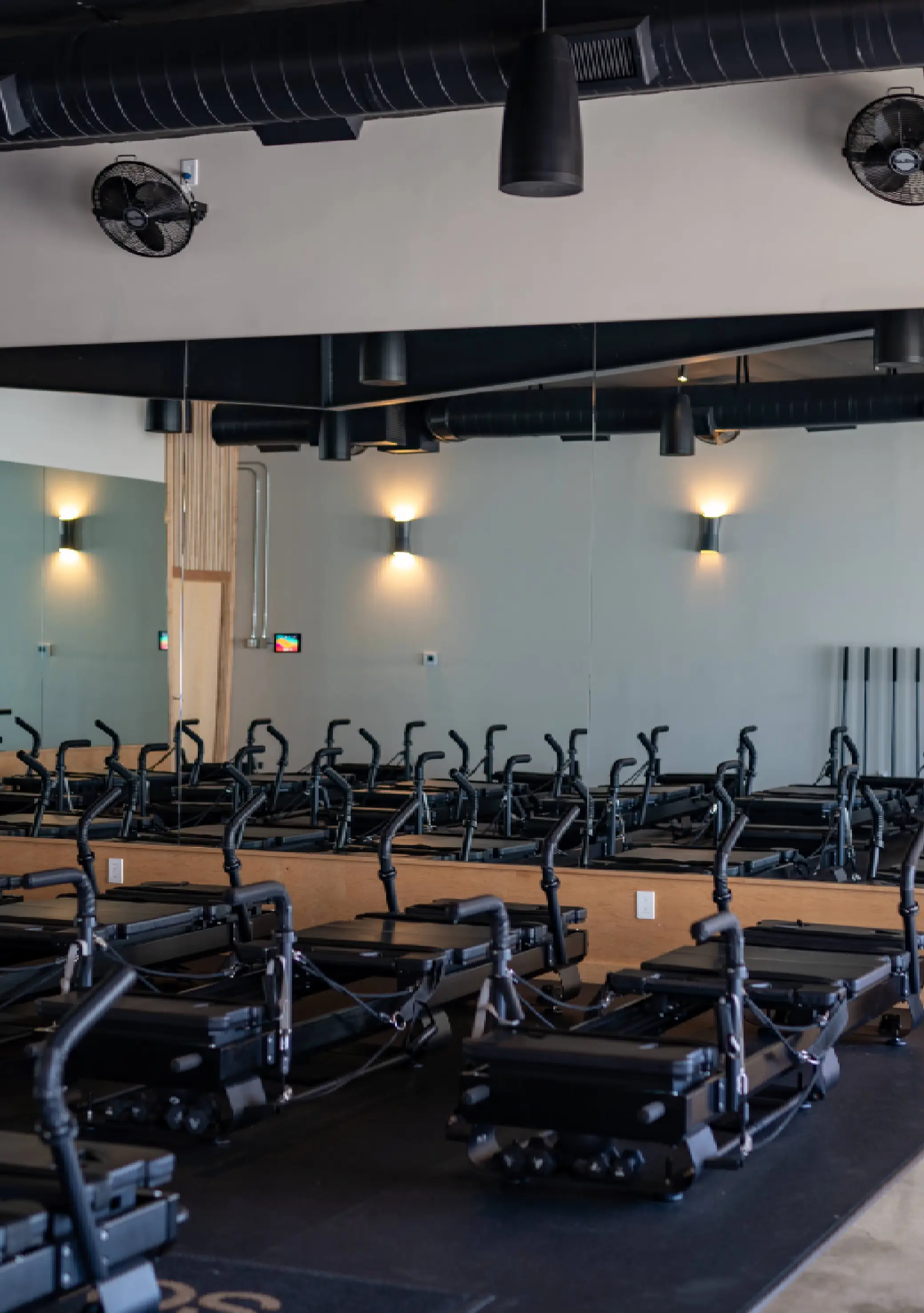 Rows of black Pilates reformer machines with handles inside a fitness studio with mirrored walls and black ceiling ducts.