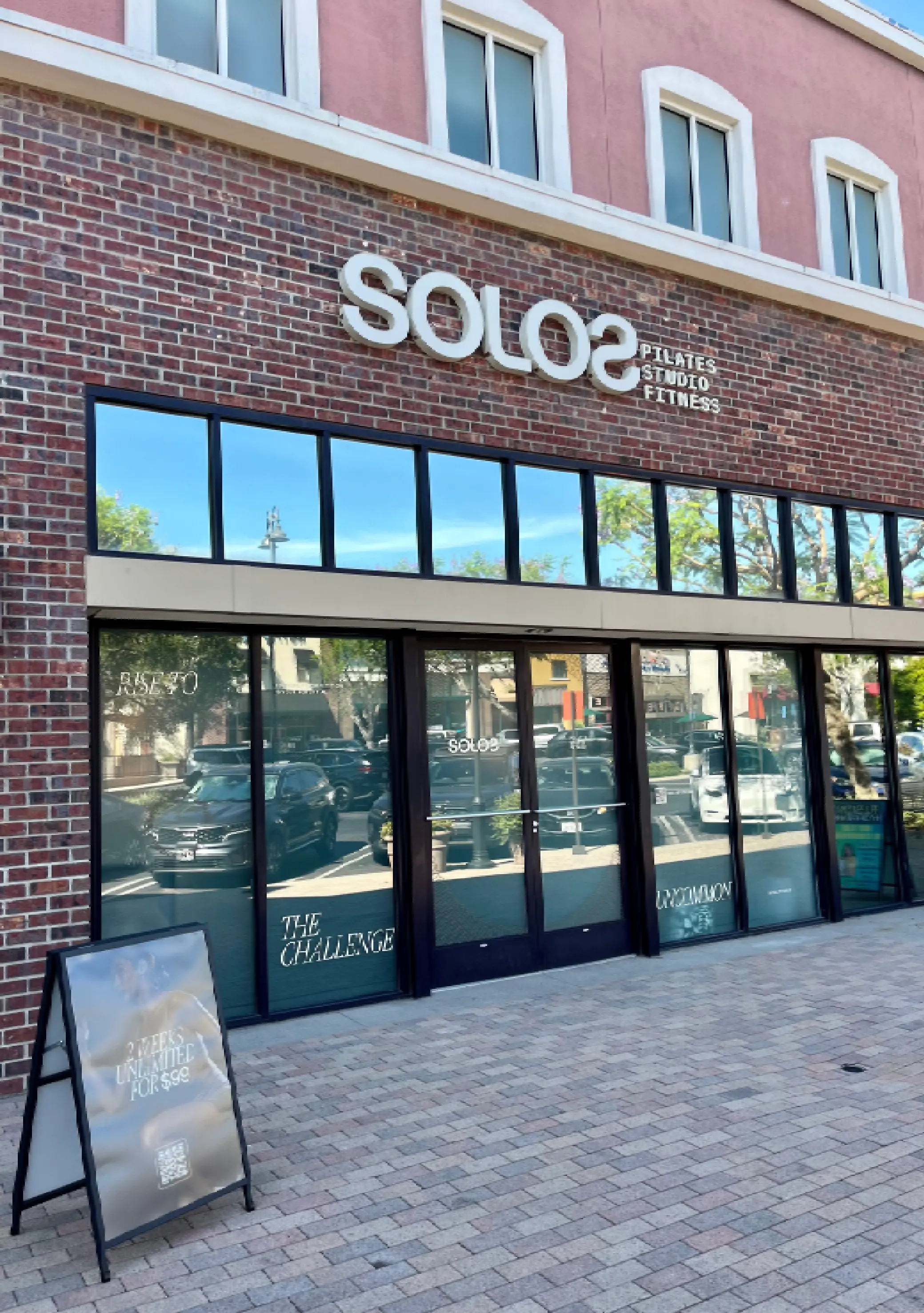 Brick building with large windows and doors, featuring 'SOLOS Pilates Studio Fitness' sign and a sidewalk sign advertising 2 weeks unlimited for $99.