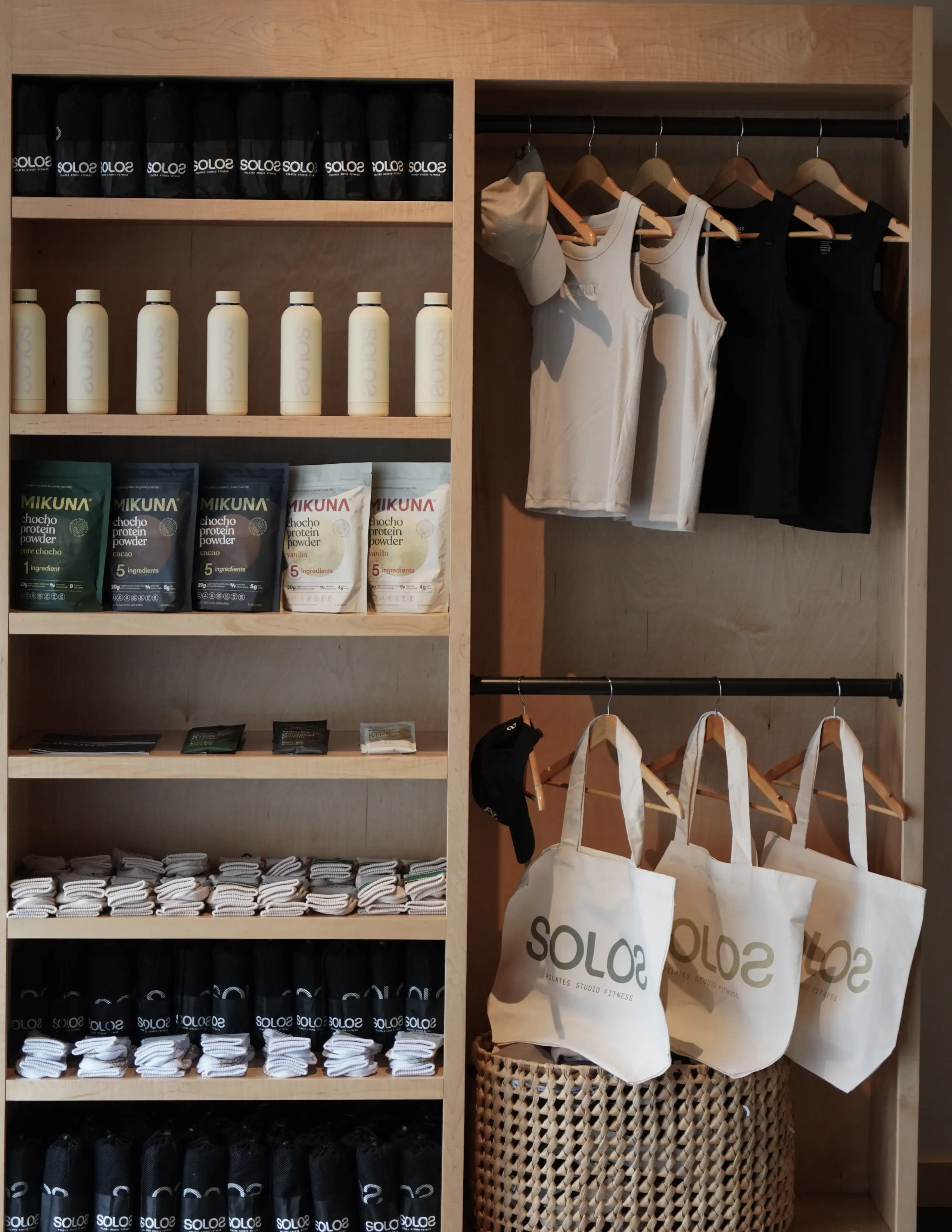 Wooden shelves and closet with fitness towels, bottles, protein powder, tank tops, caps, and tote bags.