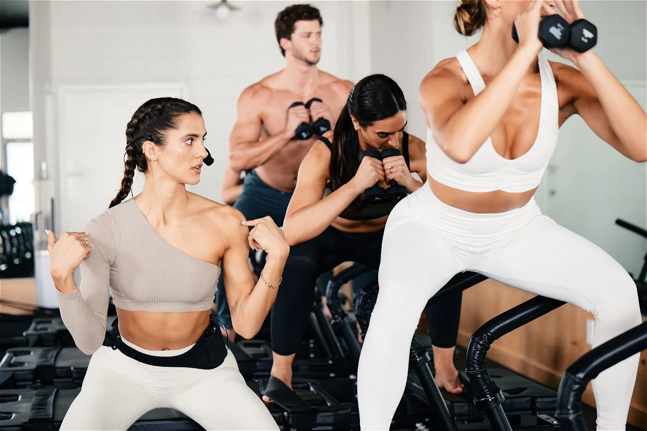 Four people exercising indoors, performing squats or lunges with dumbbells on fitness steps.