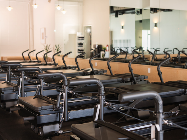 Rows of black Pilates reformer machines in a bright studio with large mirrors on the wall.