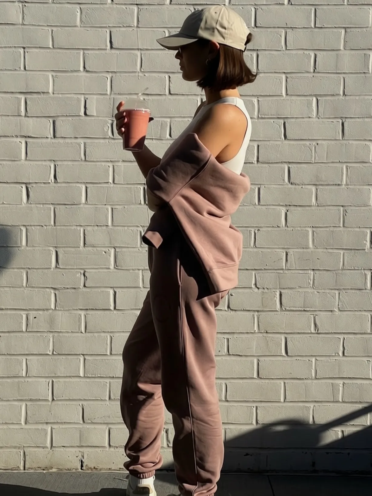 Woman in mauve sweatpants and jacket holding a pink smoothie cup standing against a white brick wall.