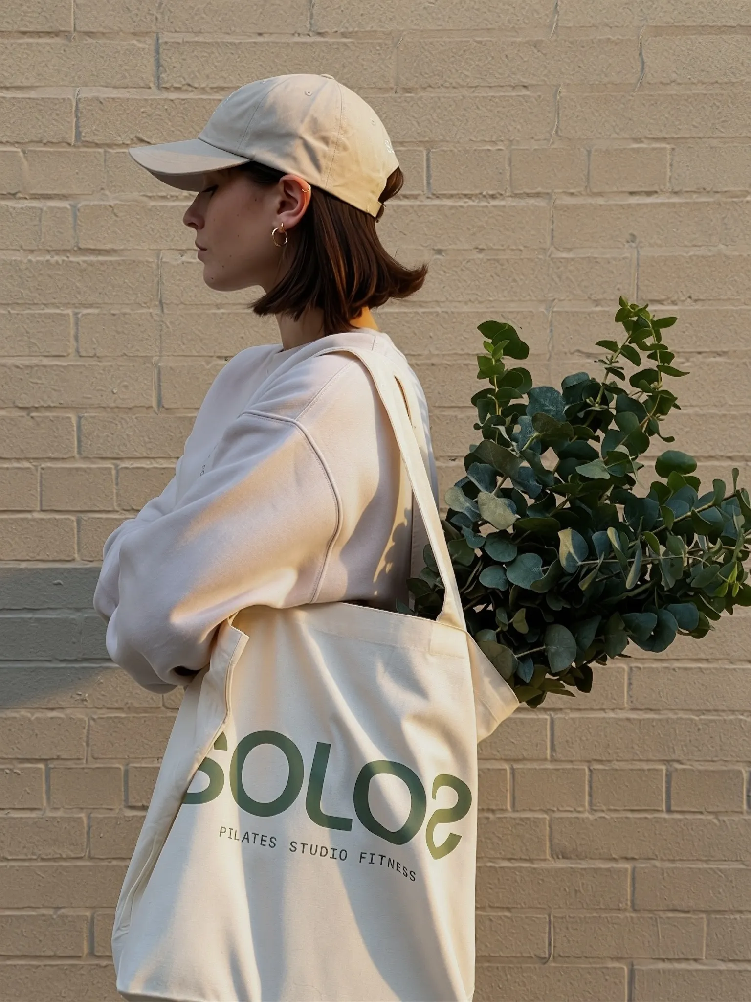 Side view of a woman in a cap holding a tote bag with green leaves against a brick wall.