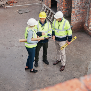 Three construction professionals in safety helmets and high-visibility vests reviewing building plans on site