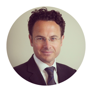 Headshot of a man with glasses, dark hair and a suit, against a grey-white background