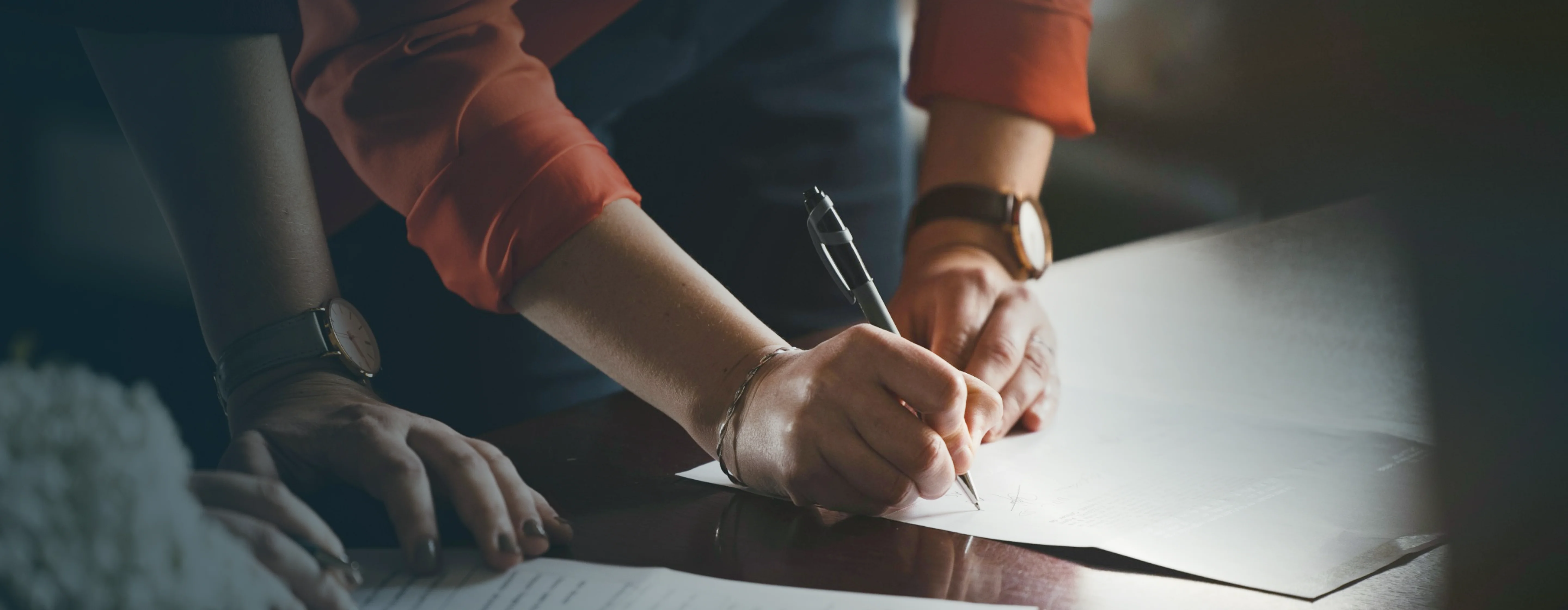 Two people, each wearing a wristwatch, reviewing and signing documents on a dark wooden table.