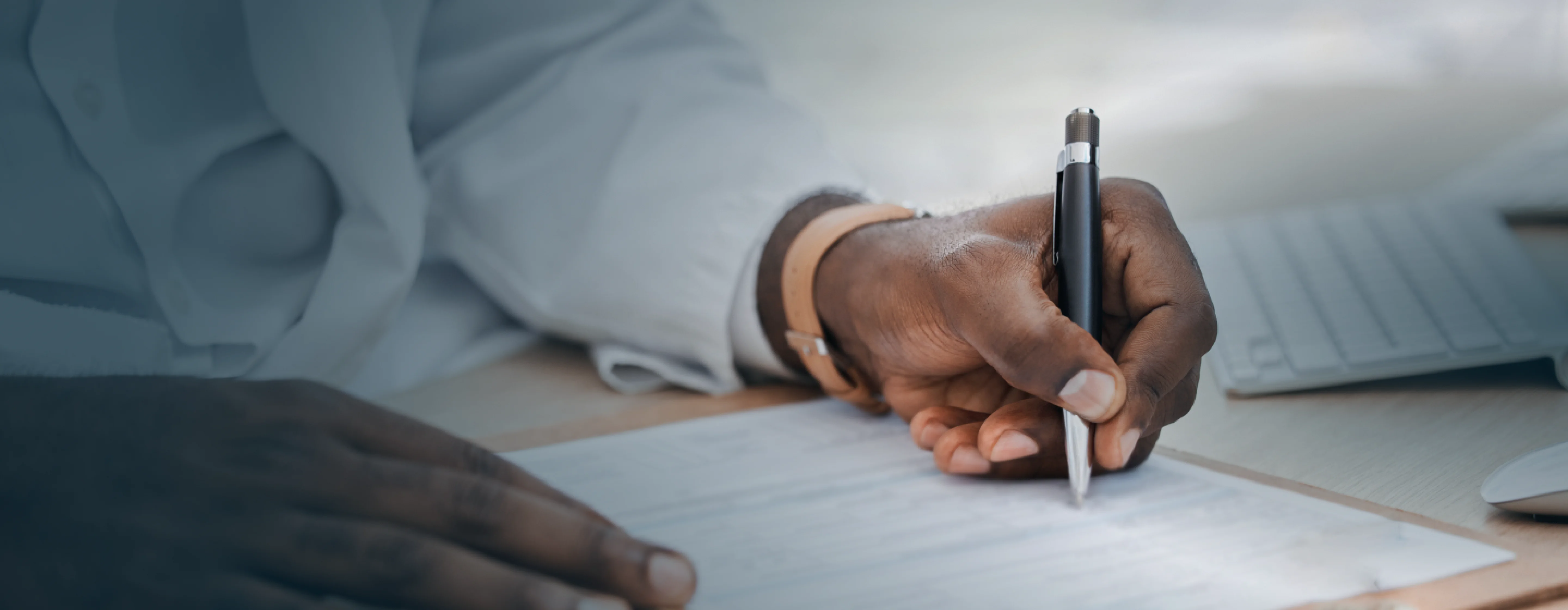 Close-up of a person writing on a document with a pen at a desk with a keyboard and mouse.