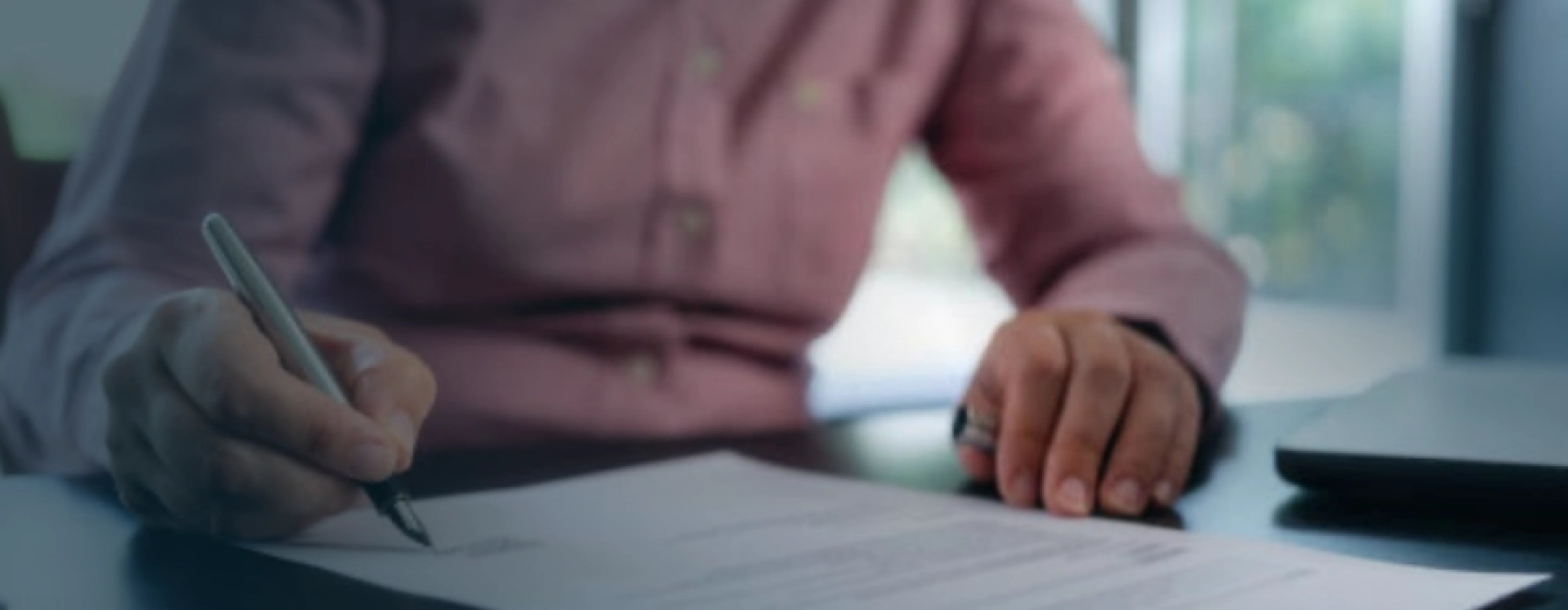 Person in a pink shirt signing a document with a pen at a desk beside a laptop.