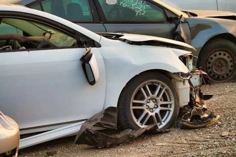 White car with severe front-end damage and detached wheel adjacent to a damaged dark-colored car.