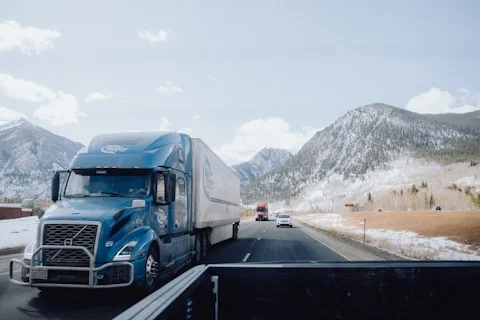 Blue semi-truck driving on a highway surrounded by snowy mountains and partly cloudy sky.