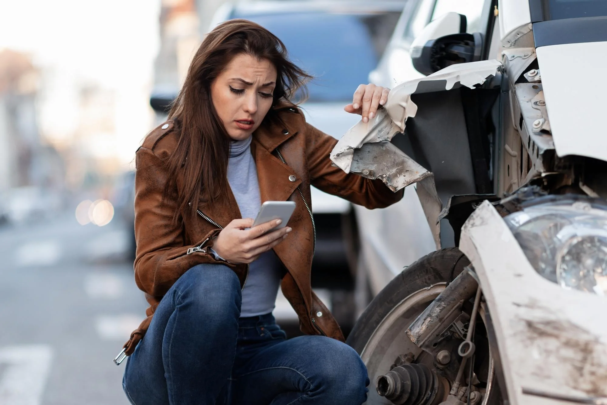 Worried woman using a smartphone while inspecting damage to a white car's front wheel and fender after an accident.