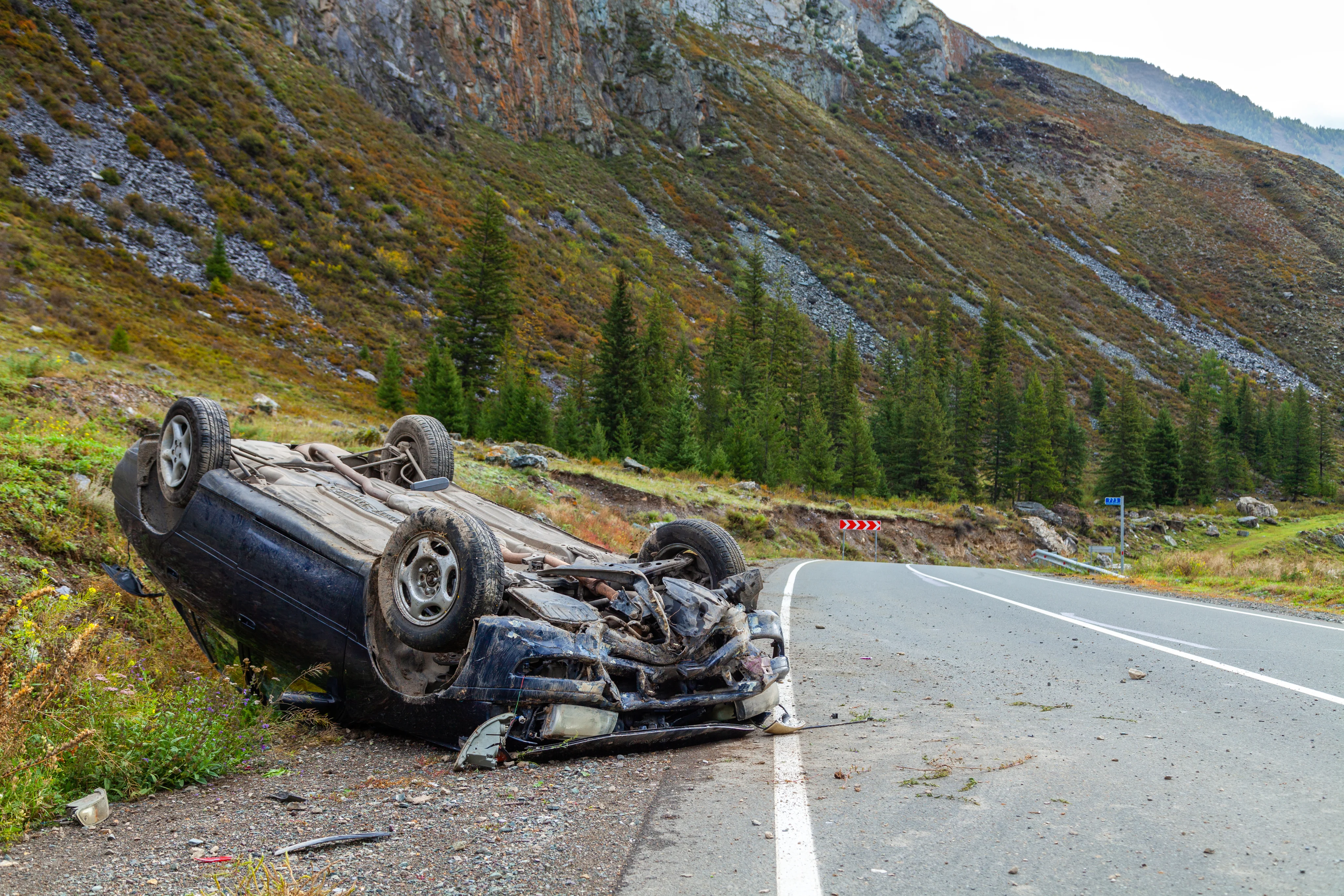 Overturned and heavily damaged car lying beside a mountain road with trees and rocky hills in the background.
