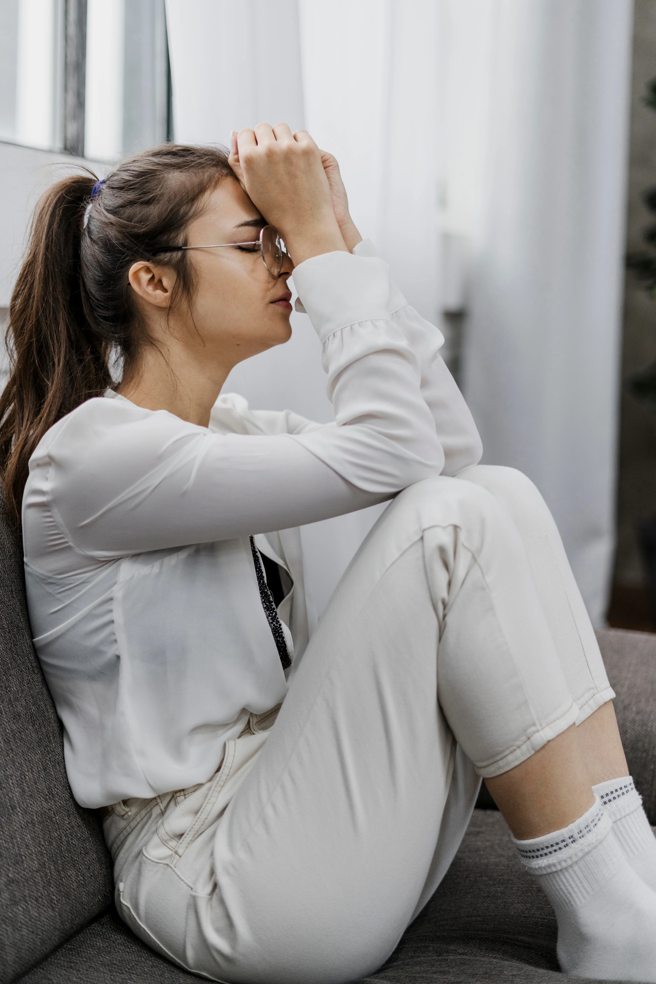 Woman wearing glasses and white clothes sits on a sofa with knees drawn up, resting her forehead on clasped hands in a contemplative pose.