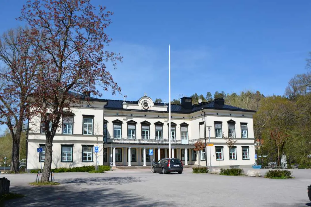 Vit byggnad i Åby med flaggstänger, grönska och mörkt tak.